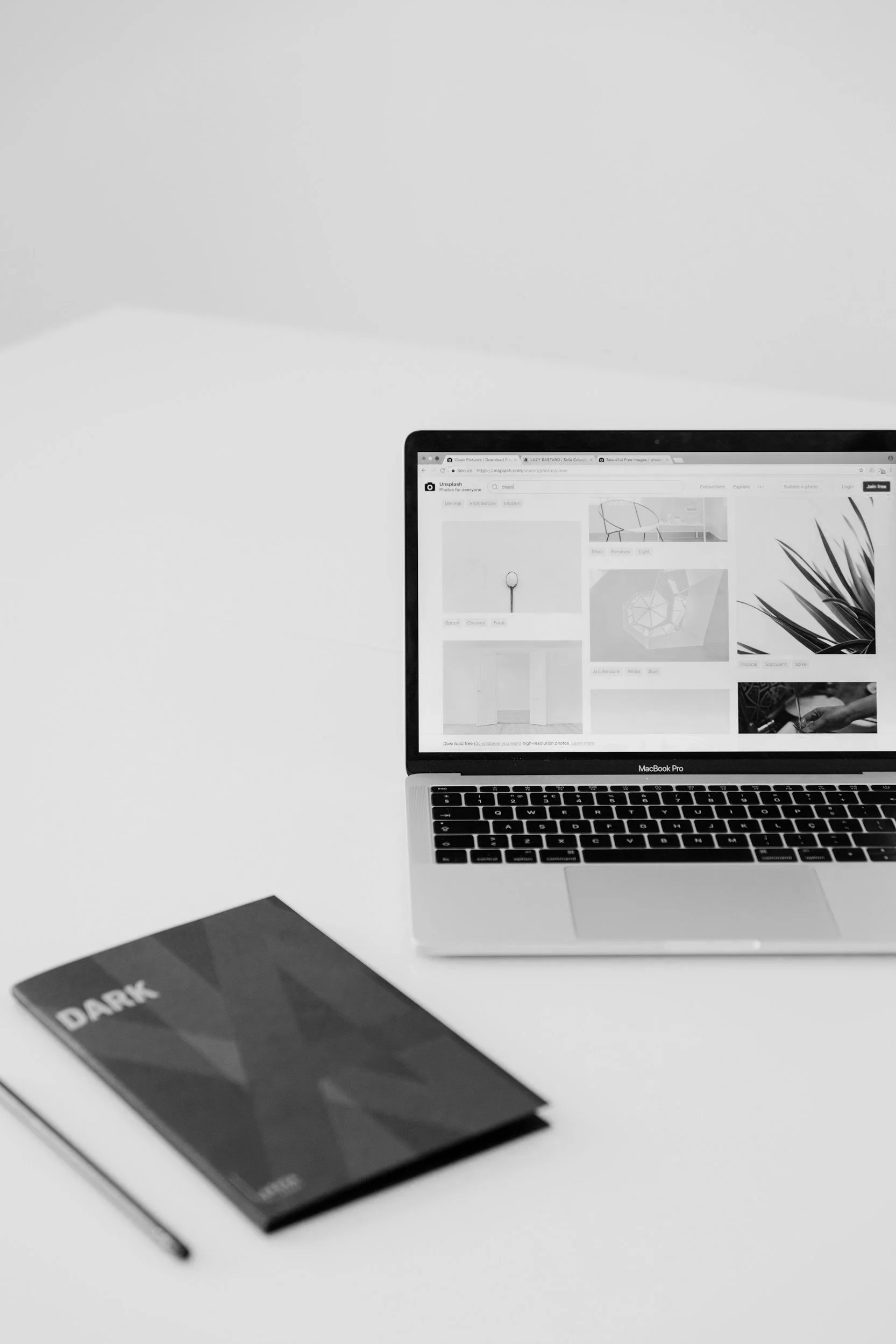 Black and white photo of a MacBook Pro laptop open on a white desk, displaying a website with images, next to a closed dark notebook labeled "DARK" and a pen.