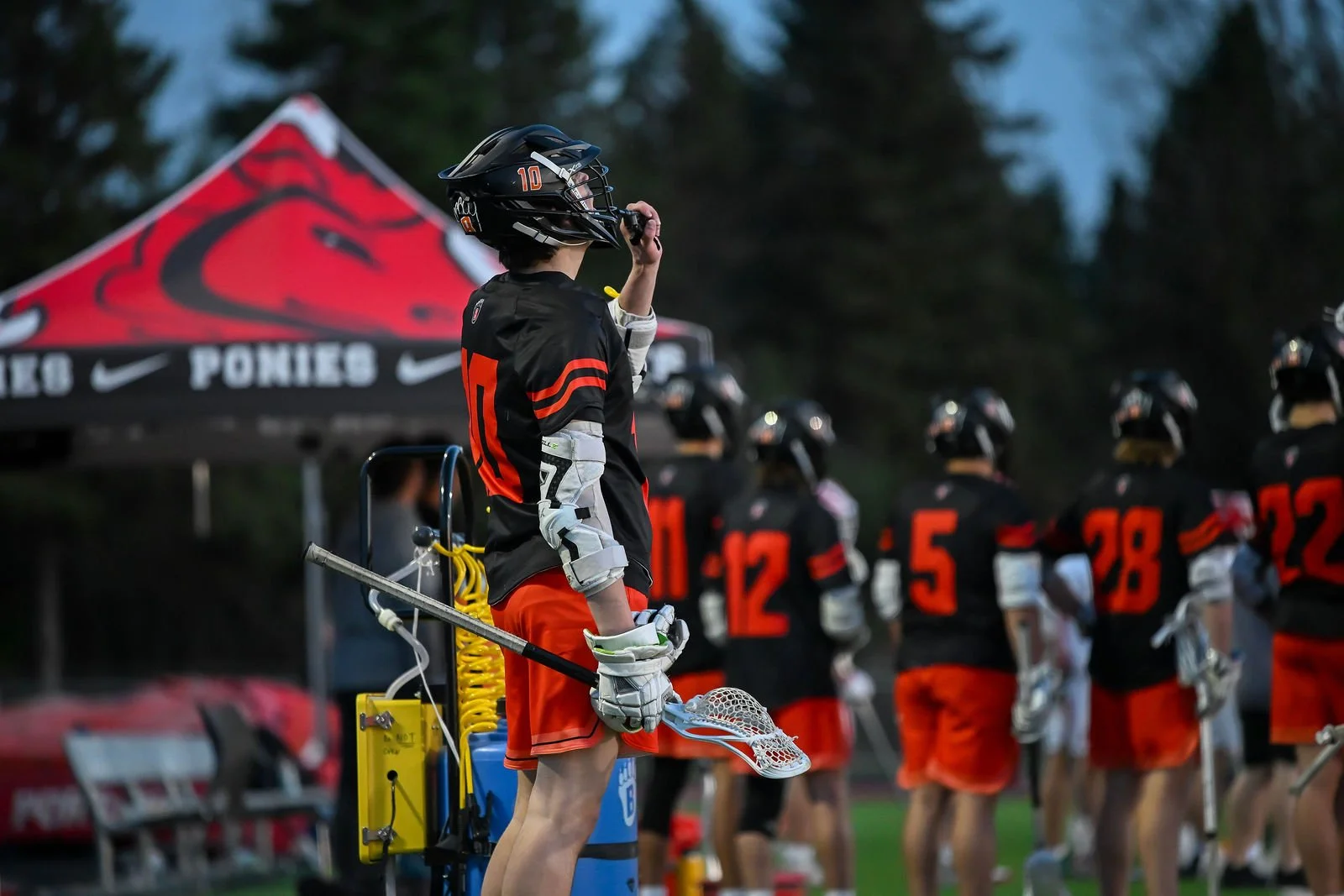 Lacrosse players in black and red jerseys, wearing helmets, standing on a field near a tent with a red logo, during evening or dusk.