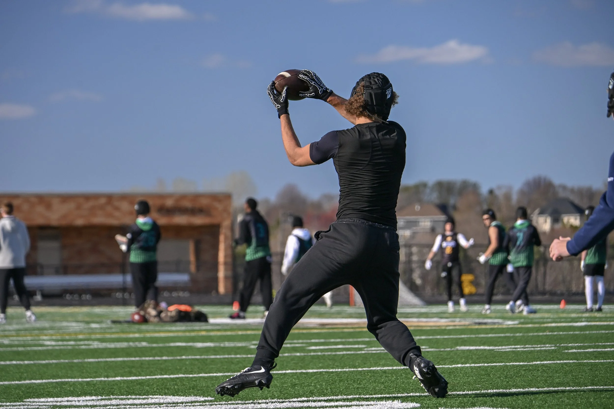 A football player wearing black athletic clothing and gloves, holding a football, preparing to throw during a game on a football field with spectators in the background.
