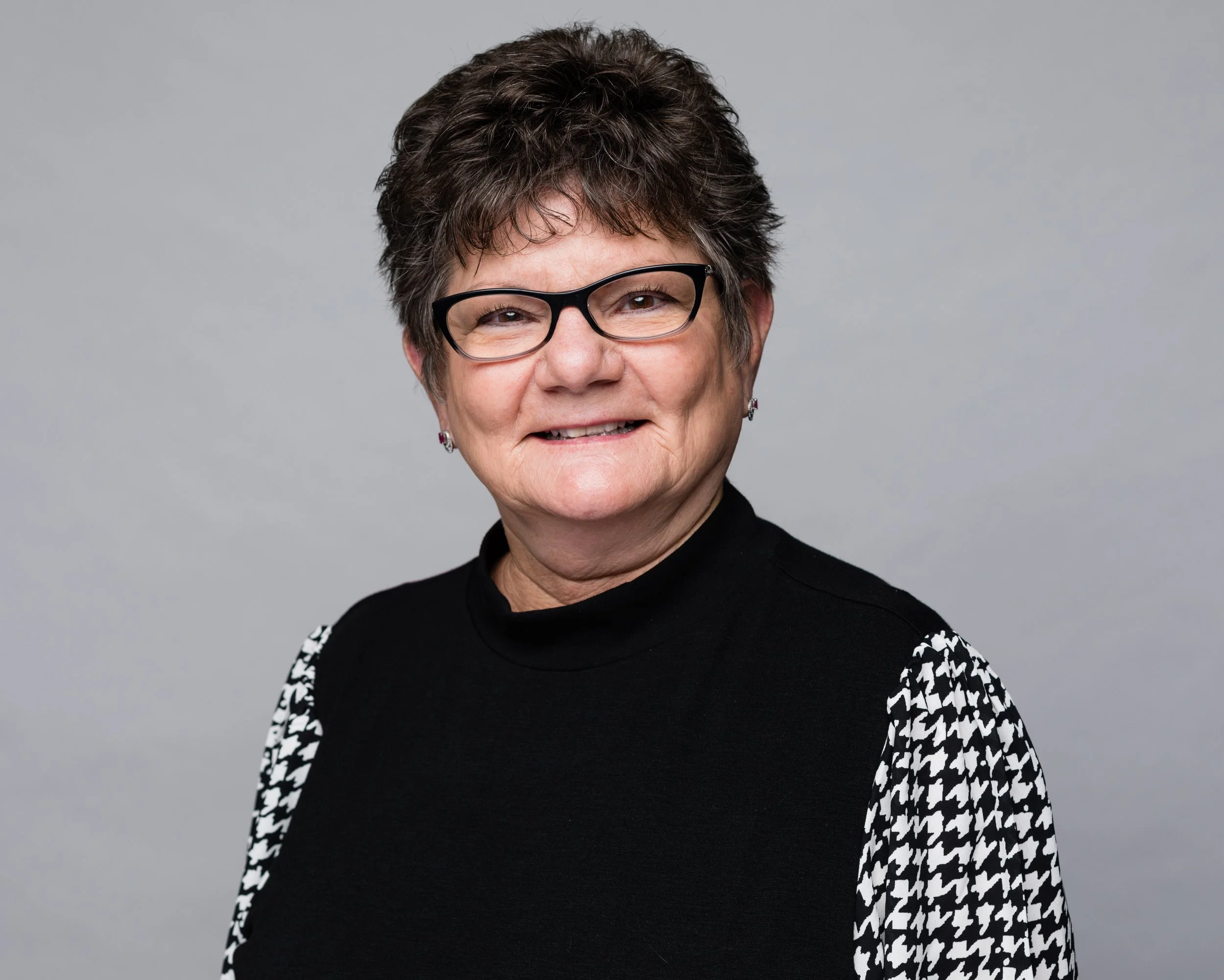 Smiling person outdoors wearing glasses and a light blue shirt, standing in front of blooming bushes, Kim Morris