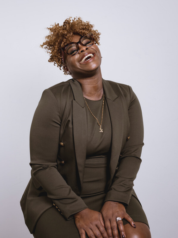 Woman with curly hair and glasses laughing joyfully, wearing an olive green blazer and dress with a gold necklace, seated against a plain light background.