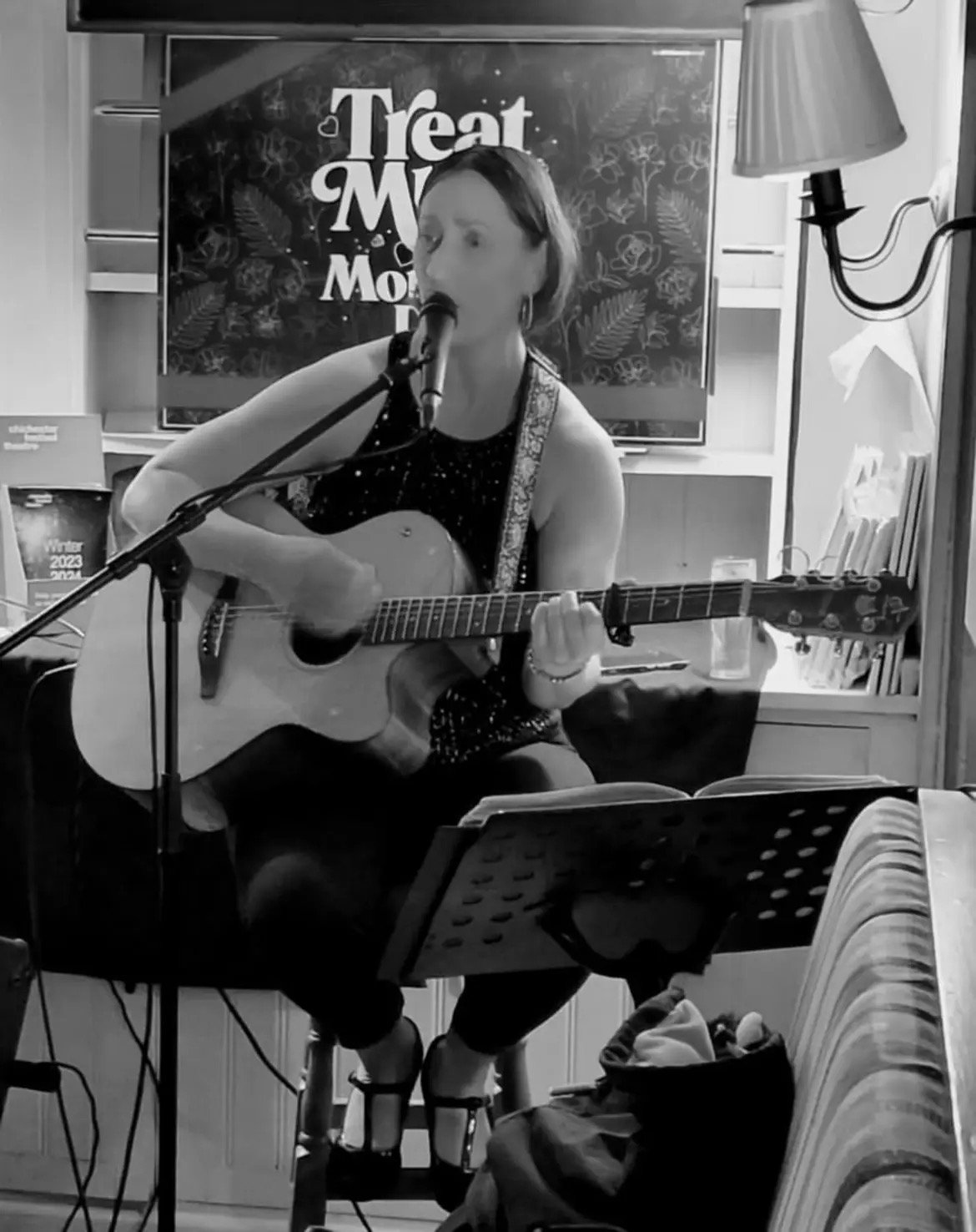 Black and white photo of a person playing an acoustic guitar and singing into a microphone. A "Treat Me" sign and music stand are visible.