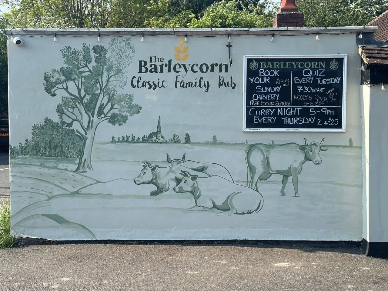 Exterior wall of The Barleycorn Classic Family Pub with mural of cows and a tree, alongside a chalkboard listing events and specials, including quiz nights and curry nights.