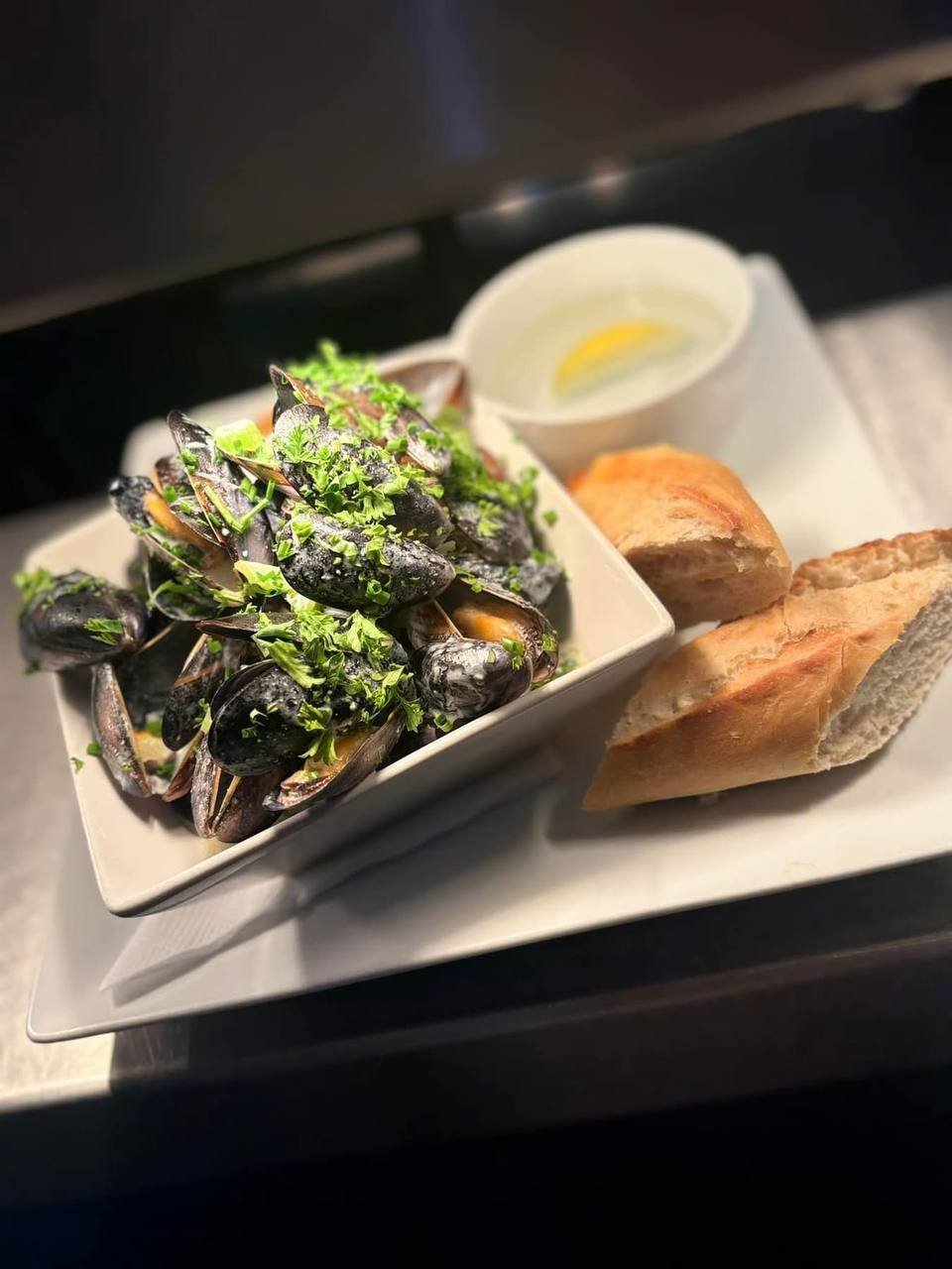 A plate of cooked mussels garnished with parsley, served with slices of baguette and a bowl of dipping sauce.