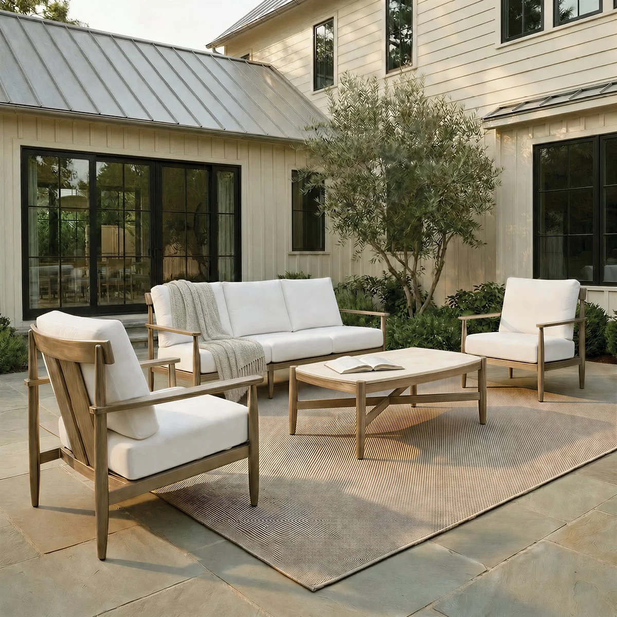 Two wicker armchairs with white cushions on a beige rug, with a small black table in between, holding a potted plant and a cup, outdoors in a garden setting with lush greenery and blue sky.