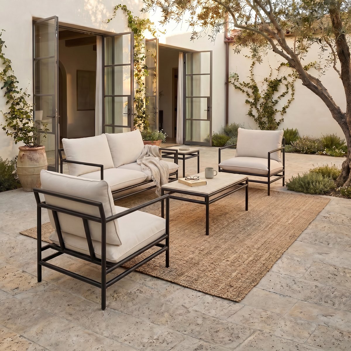 Outdoor patio with beige cushioned seating set, black metal frames, a low beige rug, potted plants, and trees, with open glass doors leading to an indoor space.