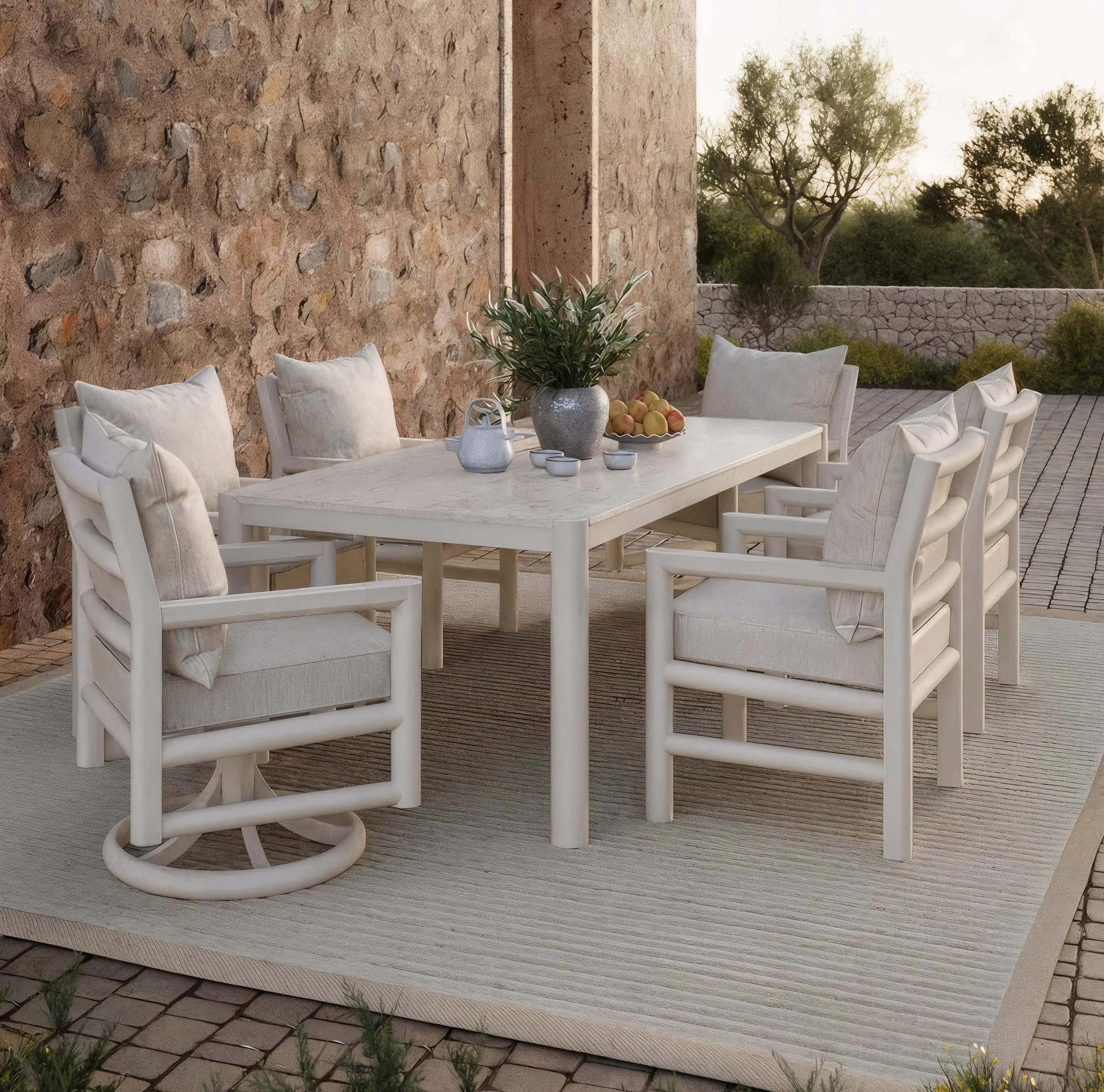 Outdoor dining area with a rectangular table, eight white cushioned chairs, a large vase with greenery, a bowl of peaches, a teapot, and cups on a stone patio in front of a brick wall and trees in the background.