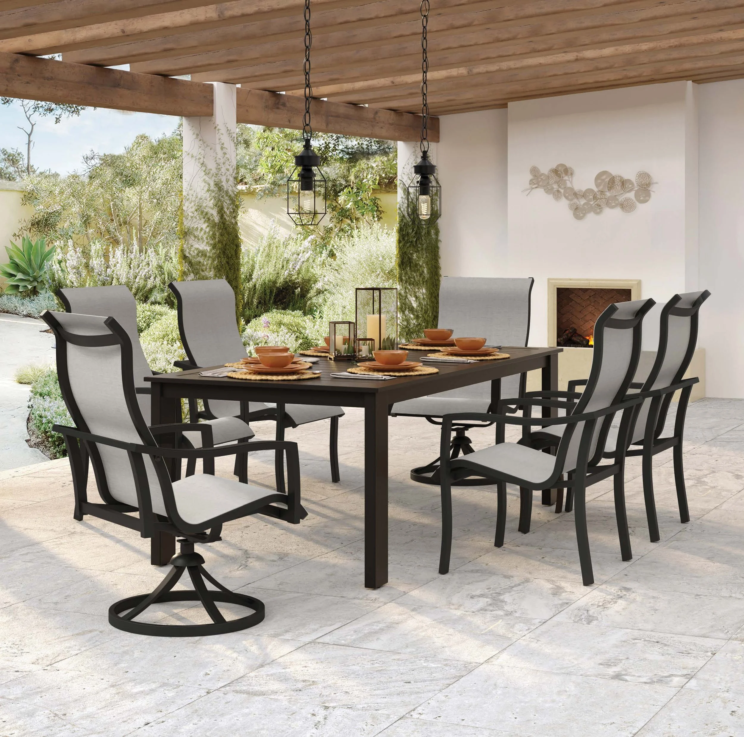 An outdoor patio with a dining table set with orange bowls, surrounded by six chairs, with a fireplace and decorative wall art, under a wooden ceiling, and lush greenery outside.