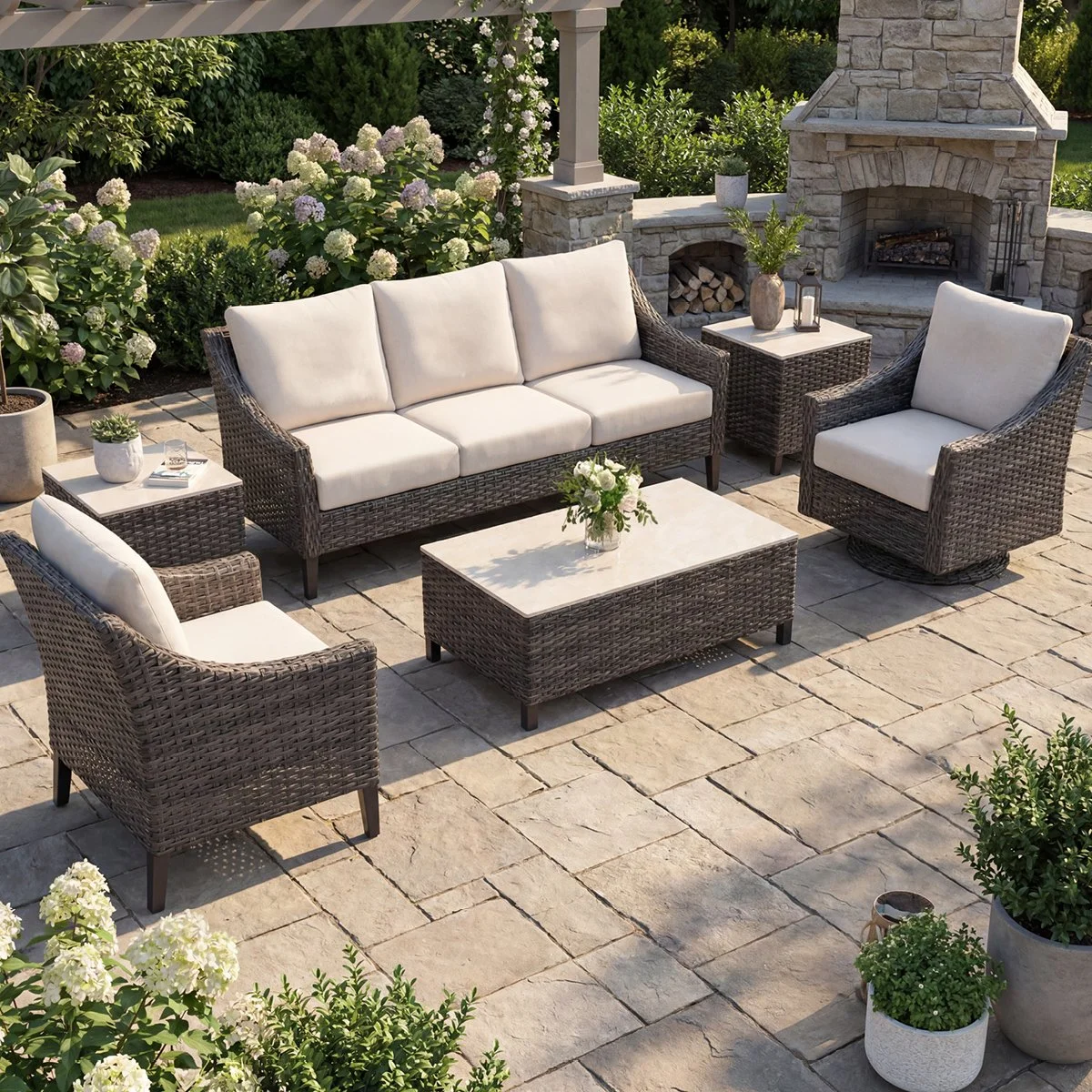 Outdoor patio with beige cushioned wicker furniture, coffee table with flowers, potted plants, a stone fireplace, and lush greenery.
