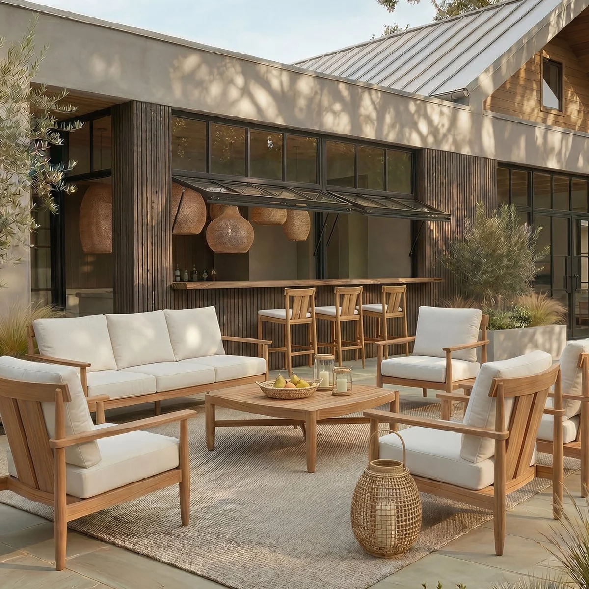 Outdoor seating area with a stone wall backdrop, featuring a white sofa with decorative pillows, two armchairs with matching pillows, a round coffee table with a bowl of fruit, a side table with a bottle and glasses, a rug, and a potted tree on the right.