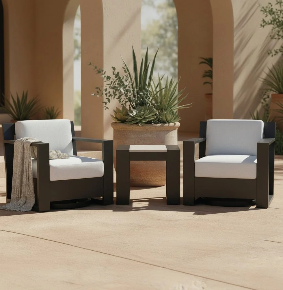 Modern outdoor patio with two cushioned chairs and a small table on a beige textured area rug. There is a house wall with large windows and a sliding glass door on the left, a white wall in the background, and some potted plants and a flowering tree on the right. Greenery is visible behind the walls.