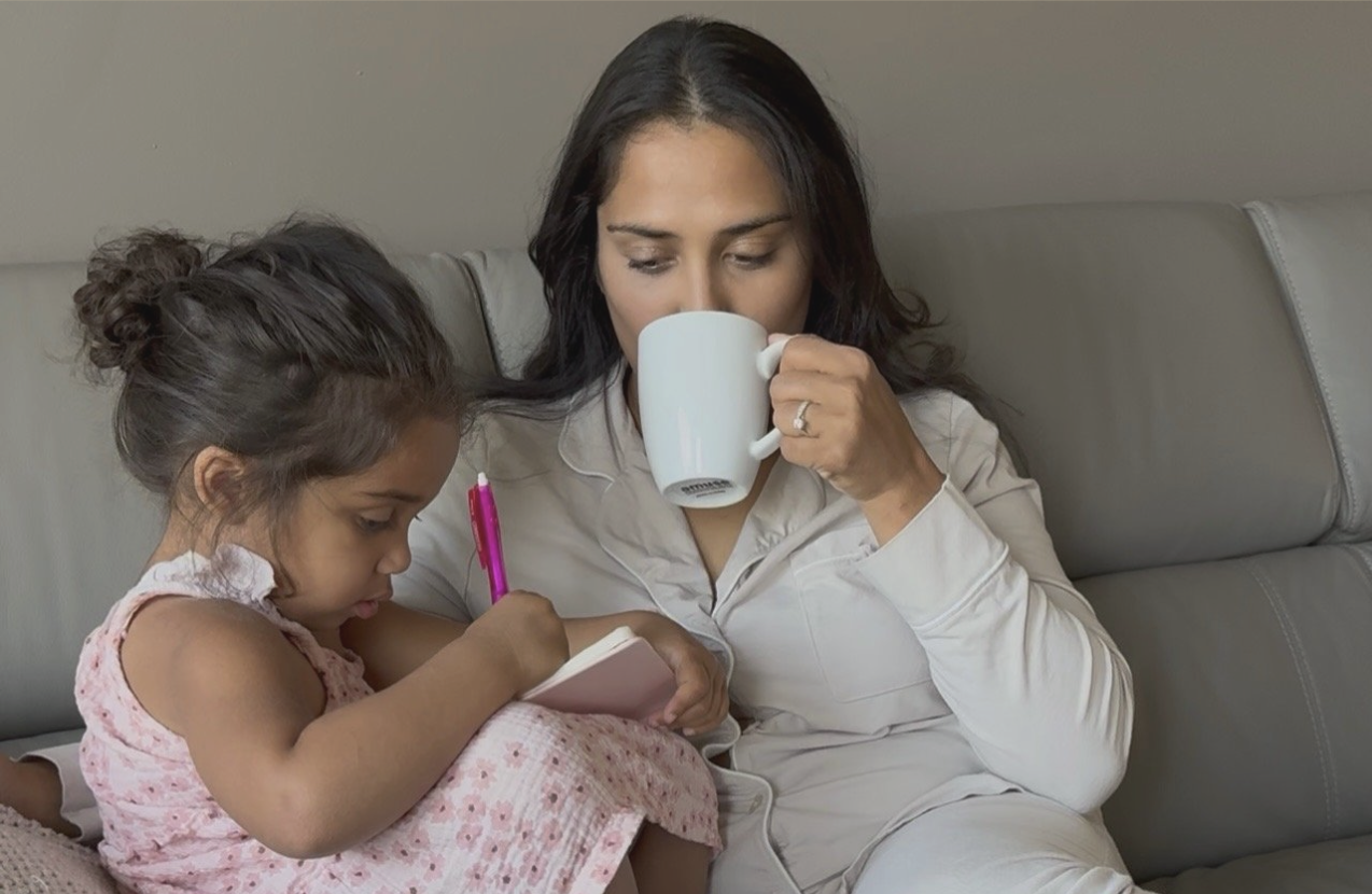 A woman with dark hair and a young girl with curly hair sitting on a gray couch, the woman drinking from a white mug while the girl sketches on a notepad.