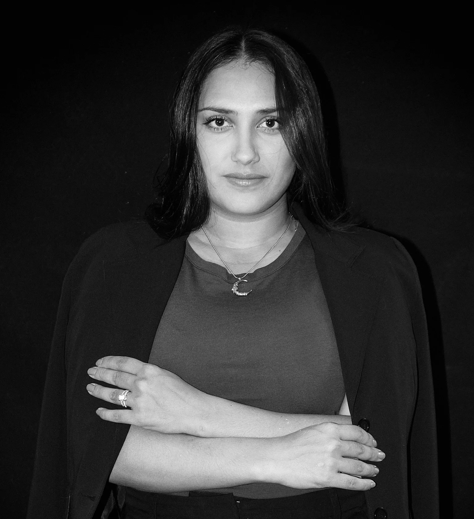 Black and white portrait of a woman with dark hair, wearing a necklace with a crescent moon pendant, a ring, and a blazer over a shirt, standing with arms crossed against a dark background.