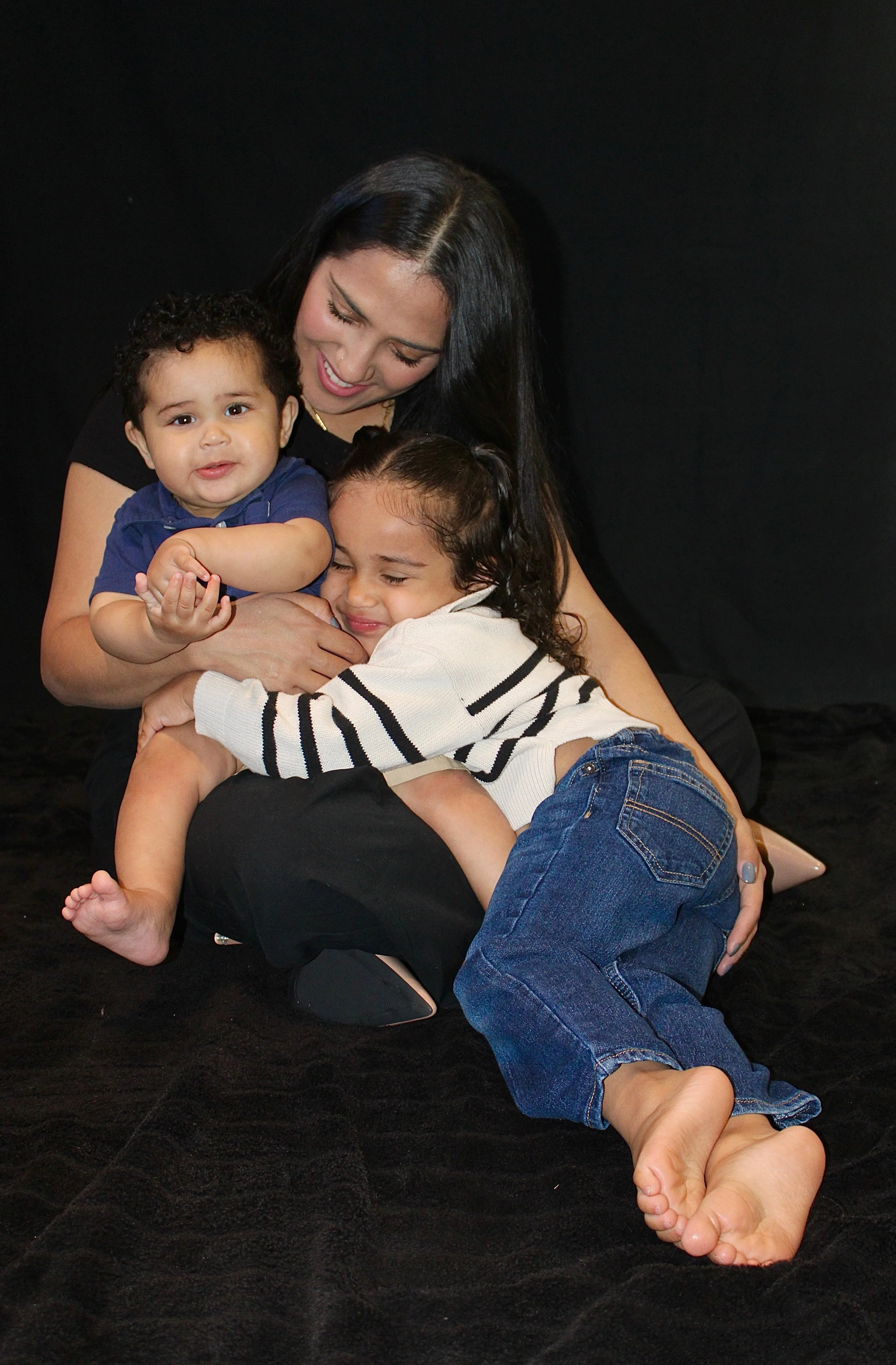 A woman with dark hair hugging two young children, a boy and a girl, against a black background. The boy has curly hair and is wearing a navy shirt, while the girl has wavy hair and is wearing a white and black striped sweater and jeans. They are all smiling and sharing a warm embrace.