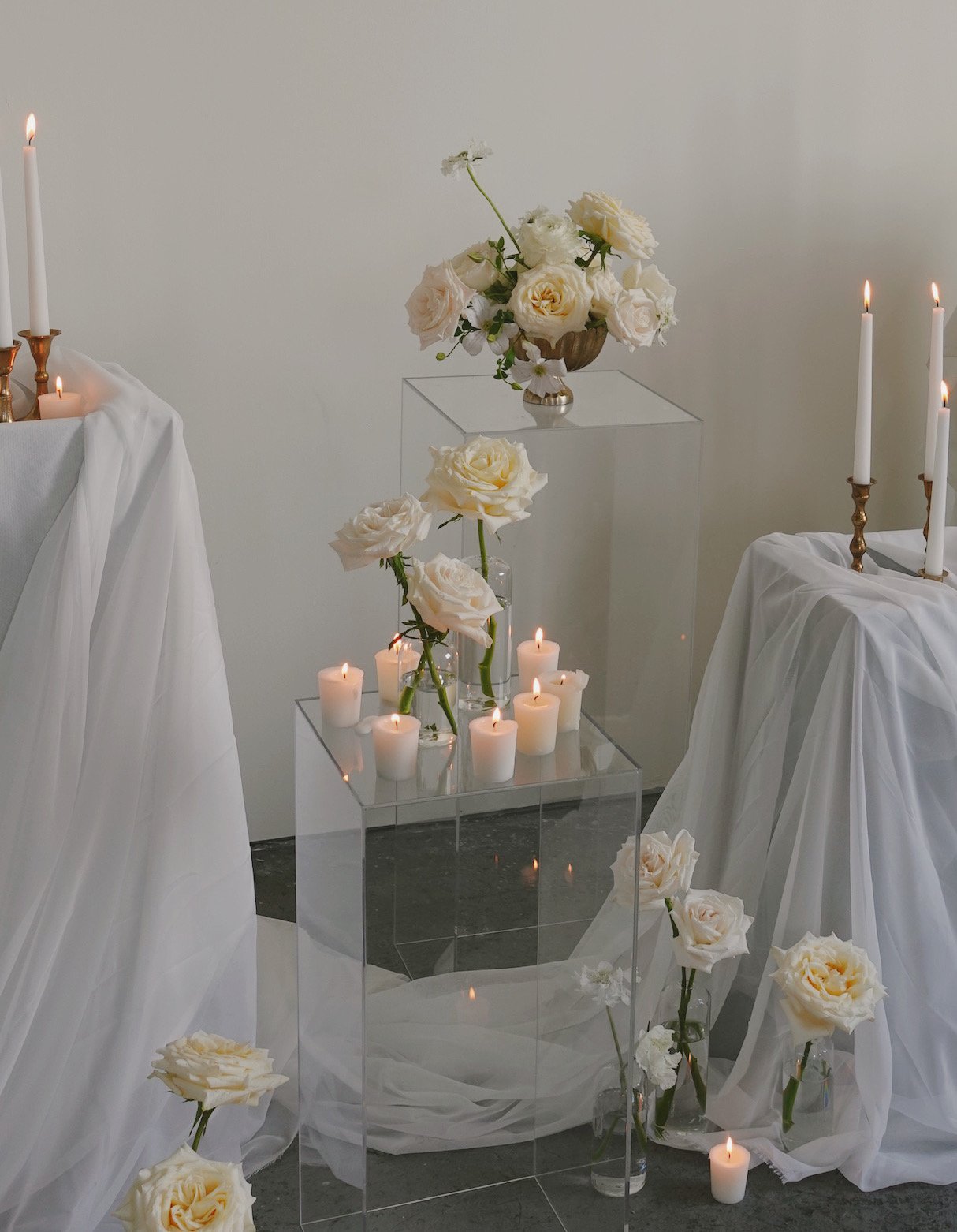 Arrangement of white roses in vases with candles on transparent stands, draped with white fabric.
