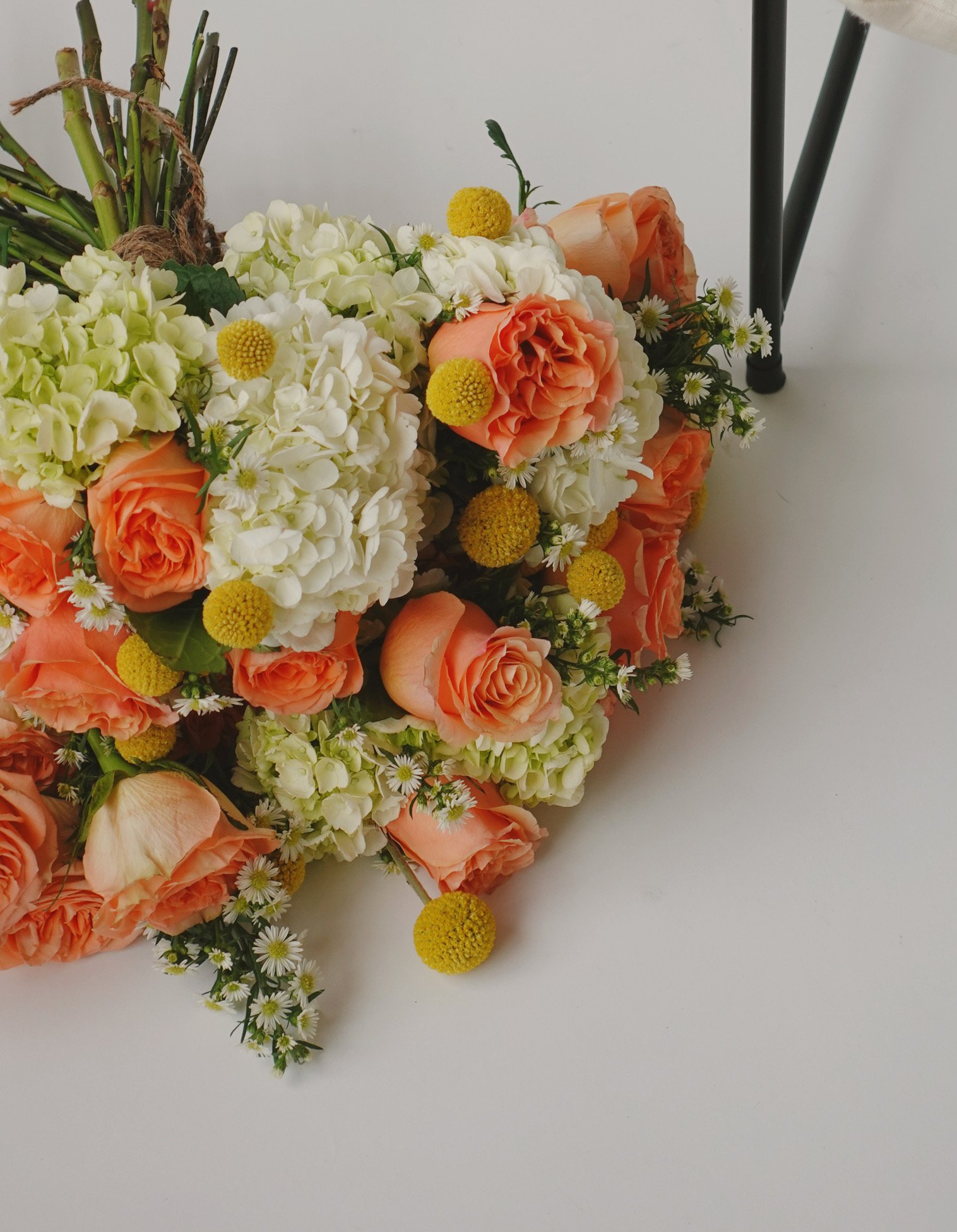 Bouquet of peach roses, white hydrangeas, yellow billy balls, and daisies on white surface.