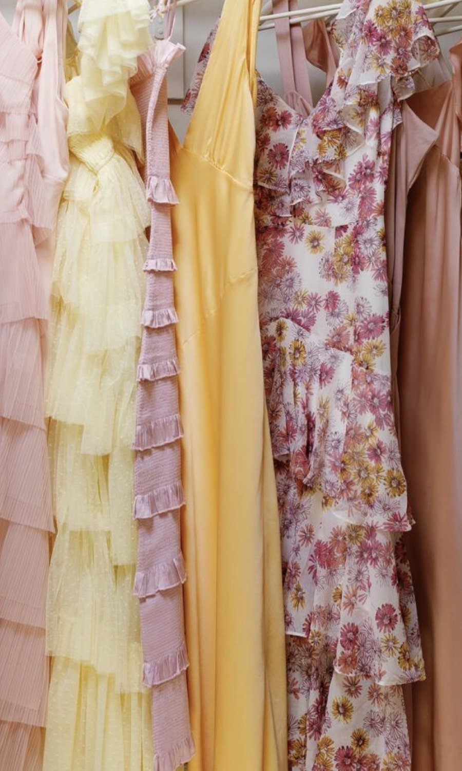 A collection of pastel and floral dresses hanging on a rack, featuring ruffles and soft colors like pink, yellow, and floral patterns.