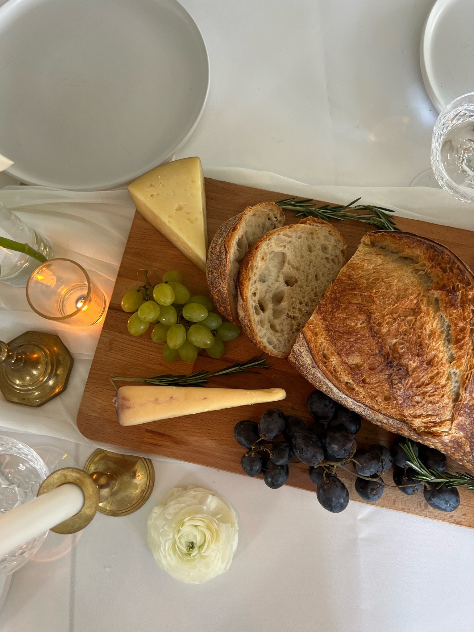 Charcuterie board with sliced bread, cheese wedges, green and black grapes, and rosemary on a wooden platter; surrounded by elegant table setting with white plates, glassware, candles, and a white flower.