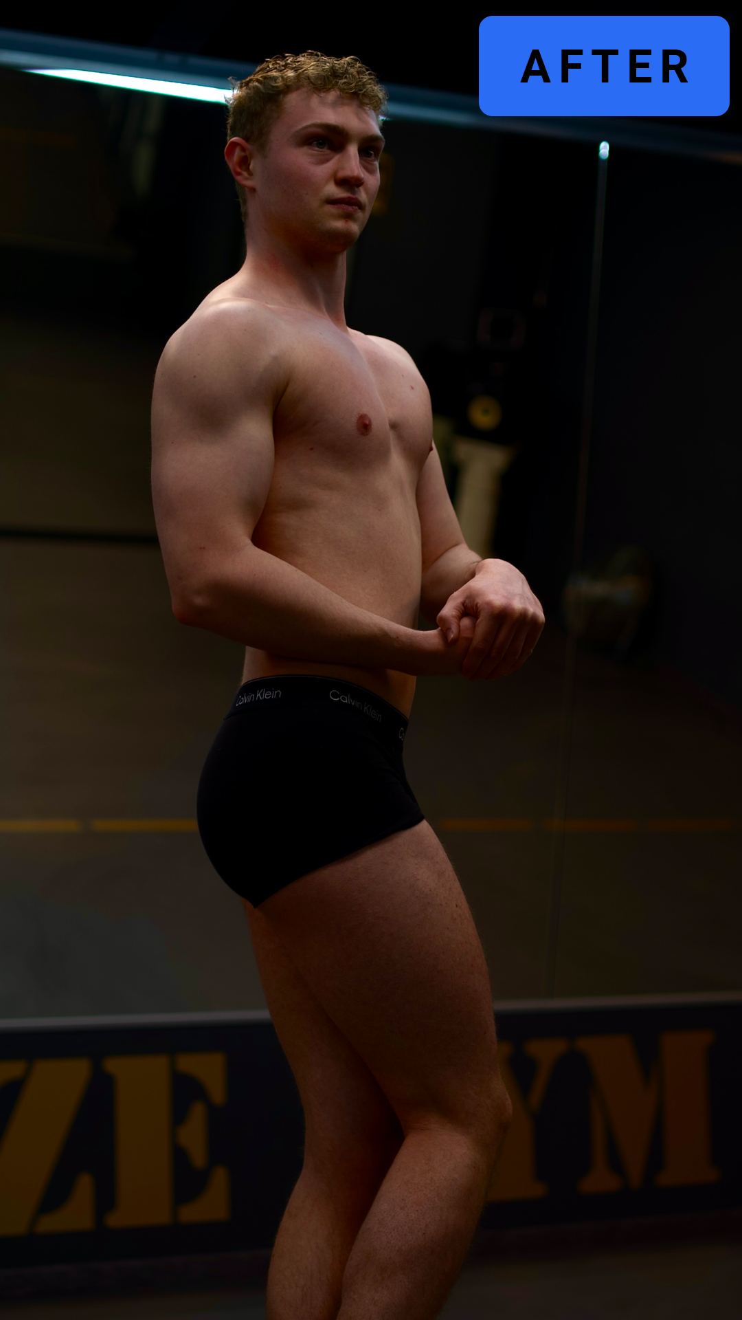 Een jonge man staat in ondergoed in een kamer met een donkere achtergrond, poserend met gevouwen handen.