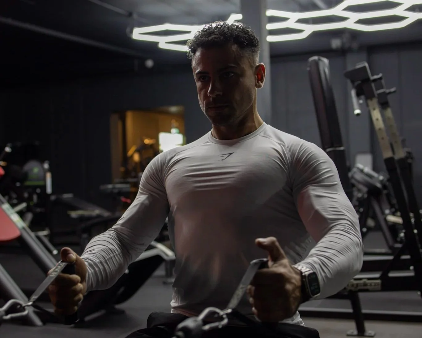 Man workout in gym, holding handles of a machine, wearing a tight white long-sleeve shirt and a smartwatch, dark background with gym equipment and modern ceiling lights.