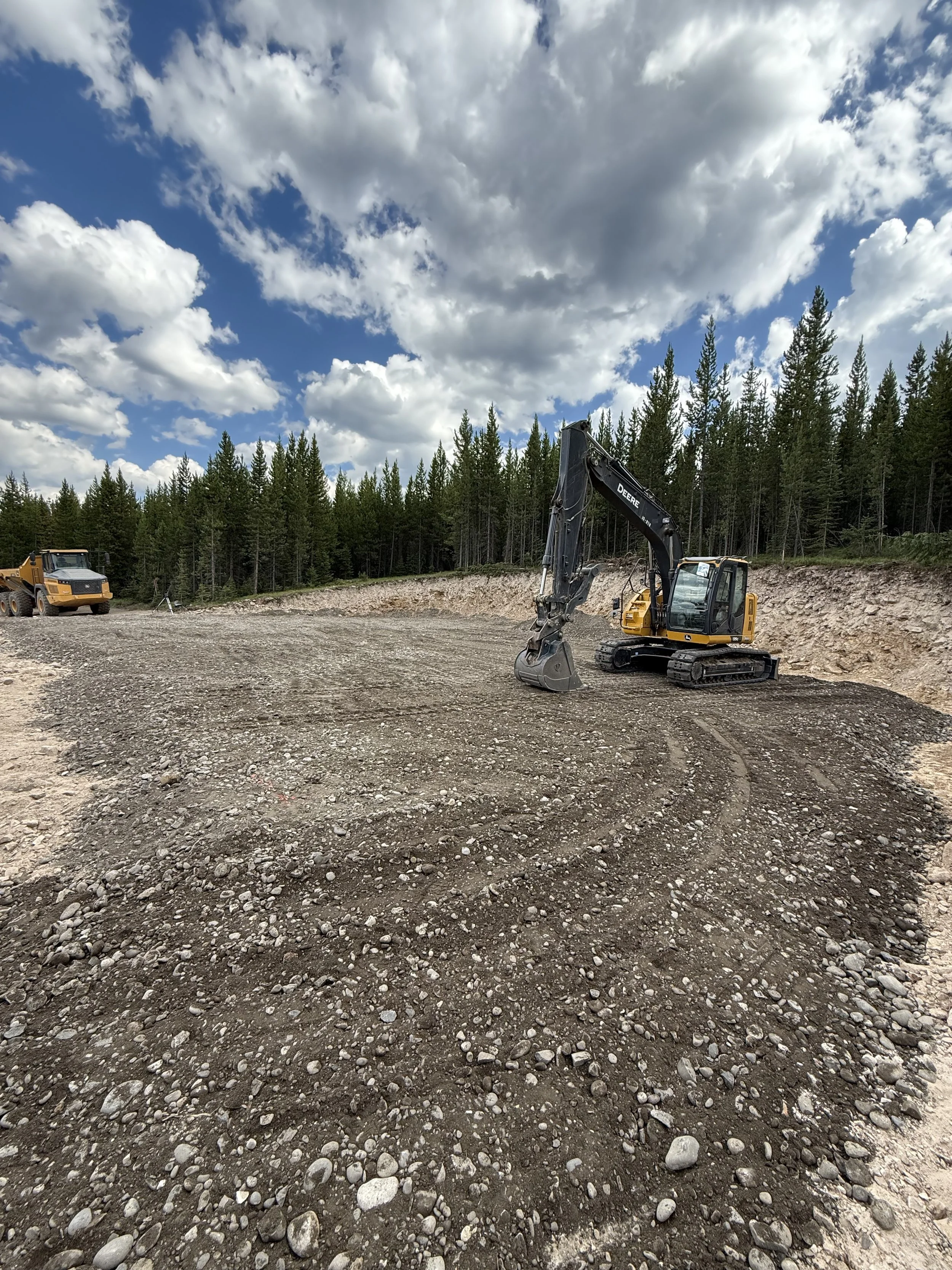 Construction site with dirt and rocks, a small excavator, and a dump truck, surrounded by a forest and covered by a partly cloudy sky.