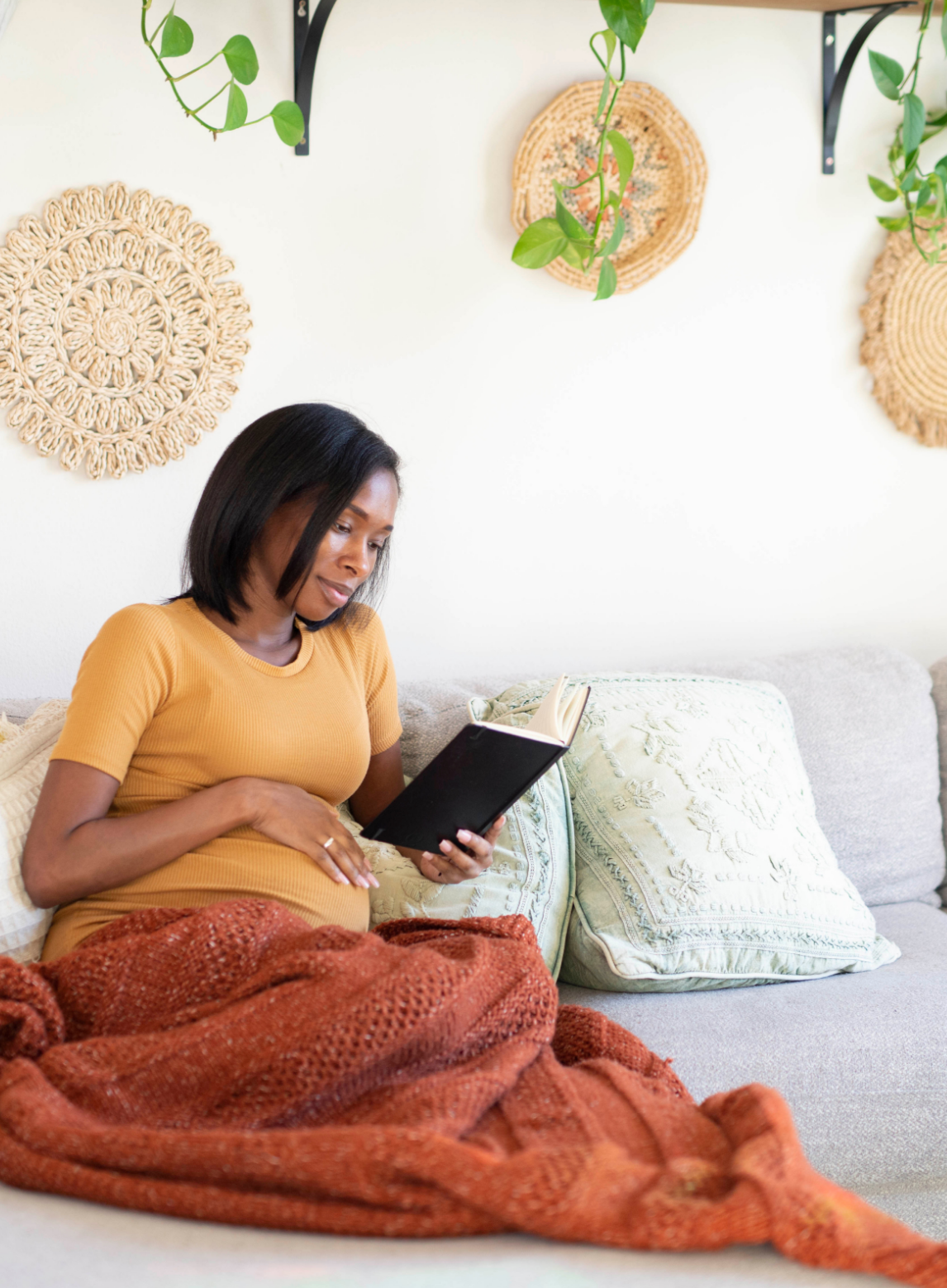 A pregnant woman sitting on a beige sofa, looking at a book, with a rust-colored blanket covering her legs. The wall behind her has woven wall hangings and hanging green plants.