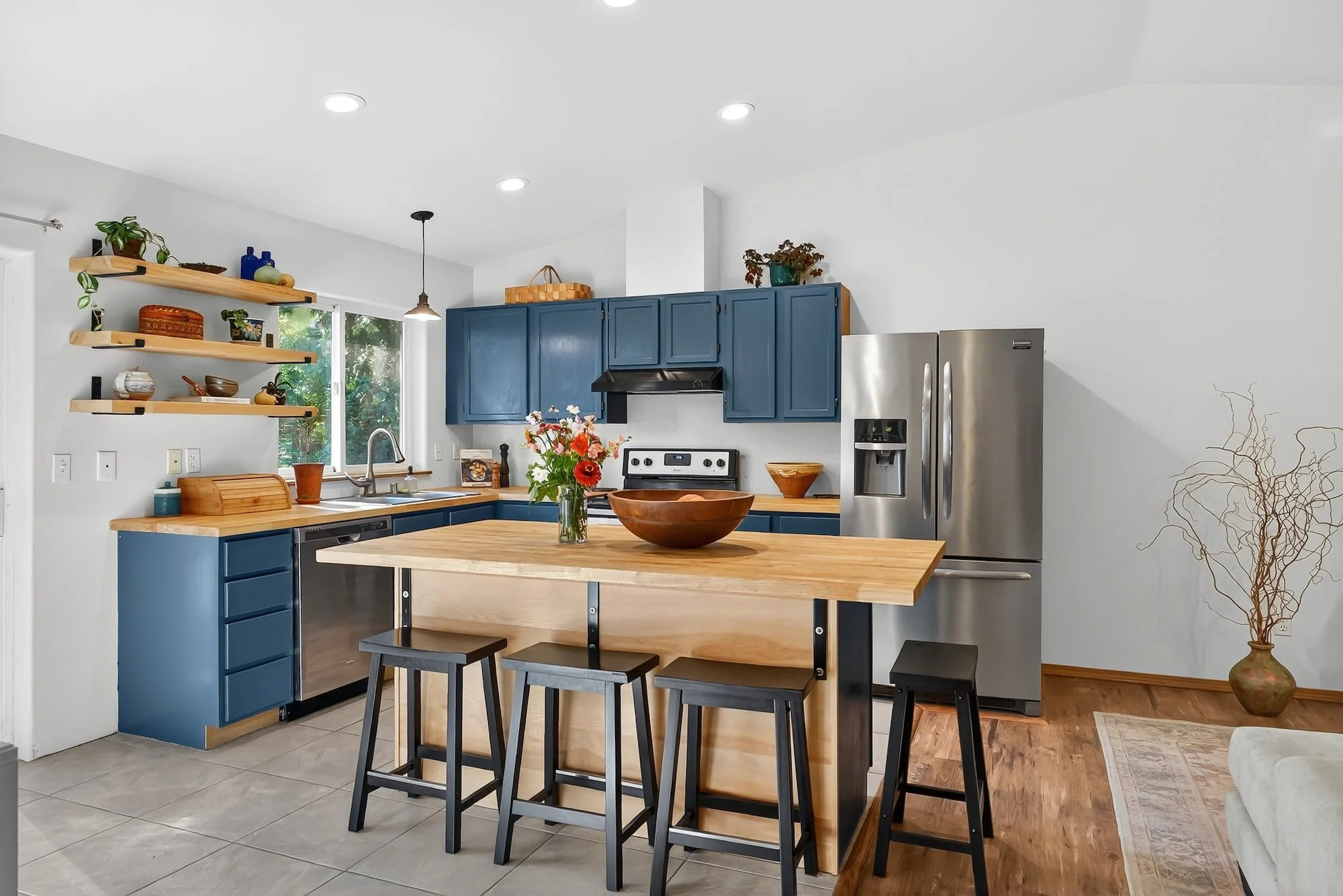 Kitchen with blue cabinets, stainless steel refrigerator, wooden island with stools, open shelving, window, and decorative items.