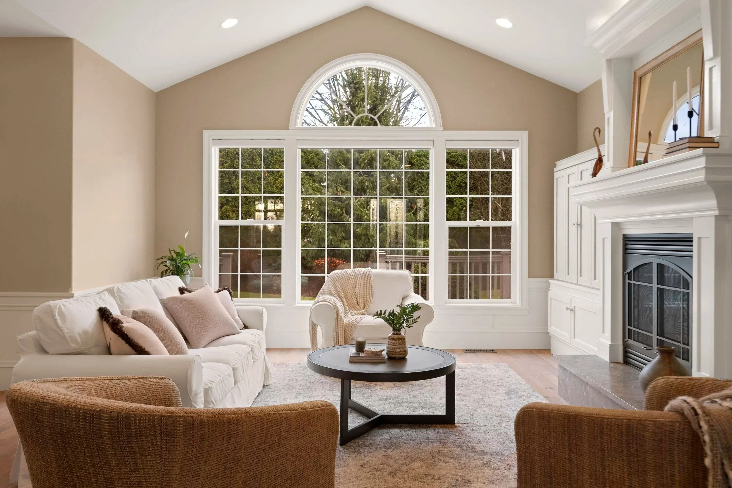 Living room with a large arched window, beige walls, white fireplace, white sofa with pink pillows, a white armchair with a blanket, two brown armchairs, a black coffee table with a plant, books, and a candle, and wooden flooring.