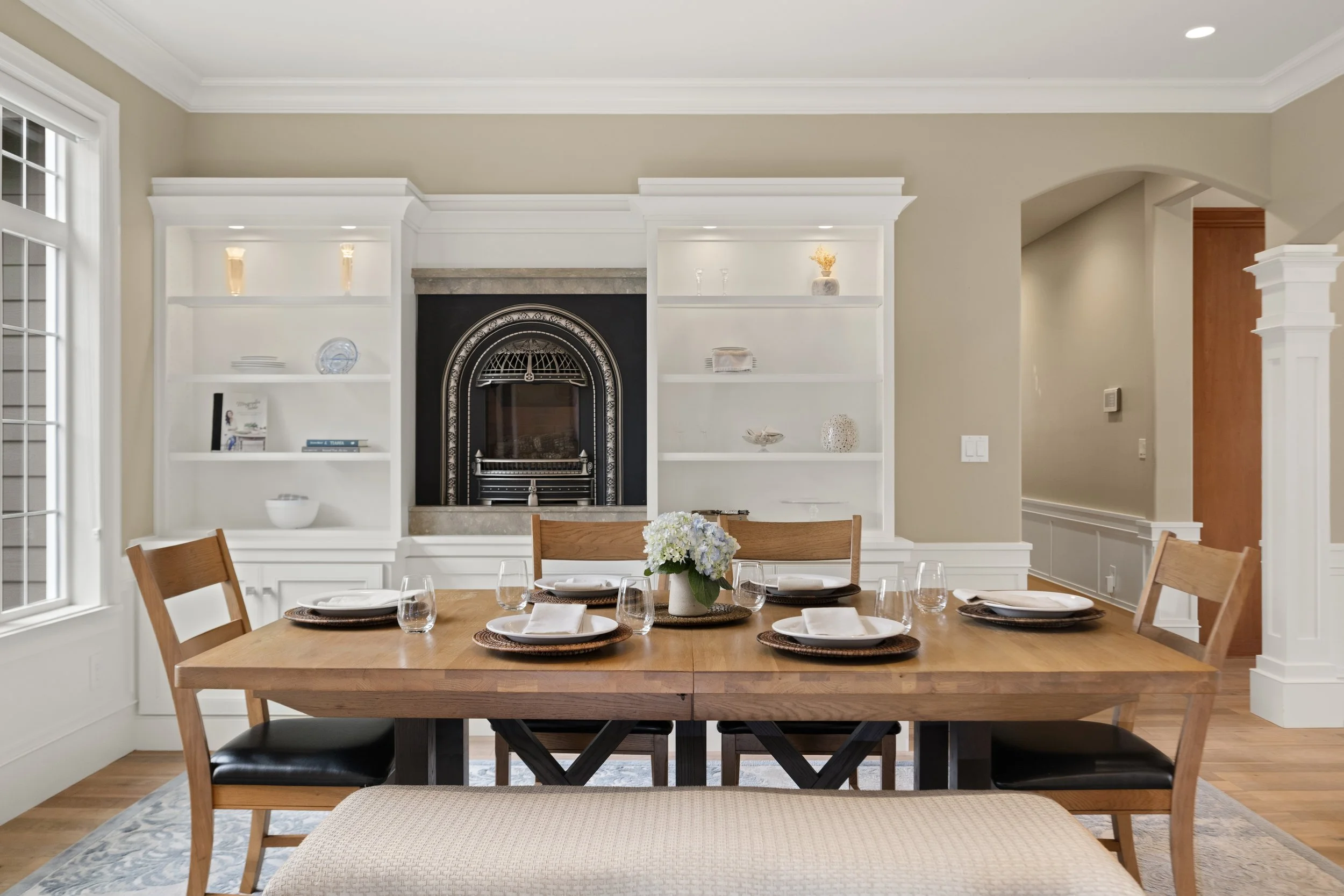 Dining room with a wooden table set with six plates, napkins, and glasses, surrounded by four wooden chairs and an upholstered bench, and a fireplace with white built-in bookshelves containing decorative items.