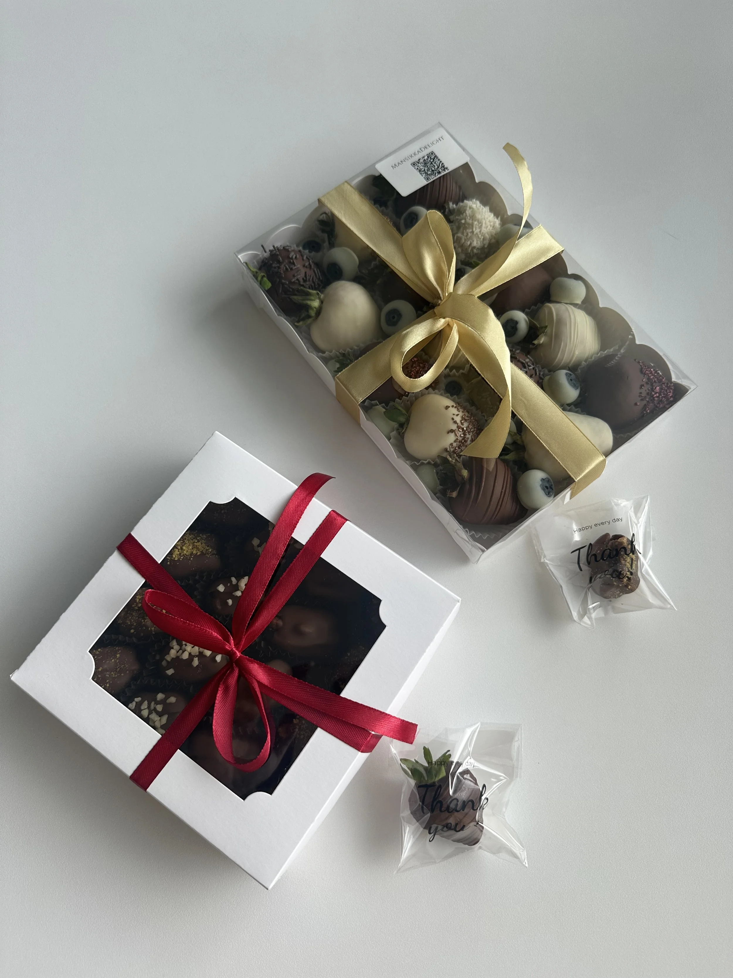 Kaksi laatikkoa ja kaksi yksittäistä suklaakarkkia, yksi valkoisessa pakkauksessa ja toinen läpinäkyvässä pakkauksessa, joka sisältää pieniä suklaamukeja. Kaikissa pakkauksissa on rusetti, toinen kultainen ja toinen punainen.