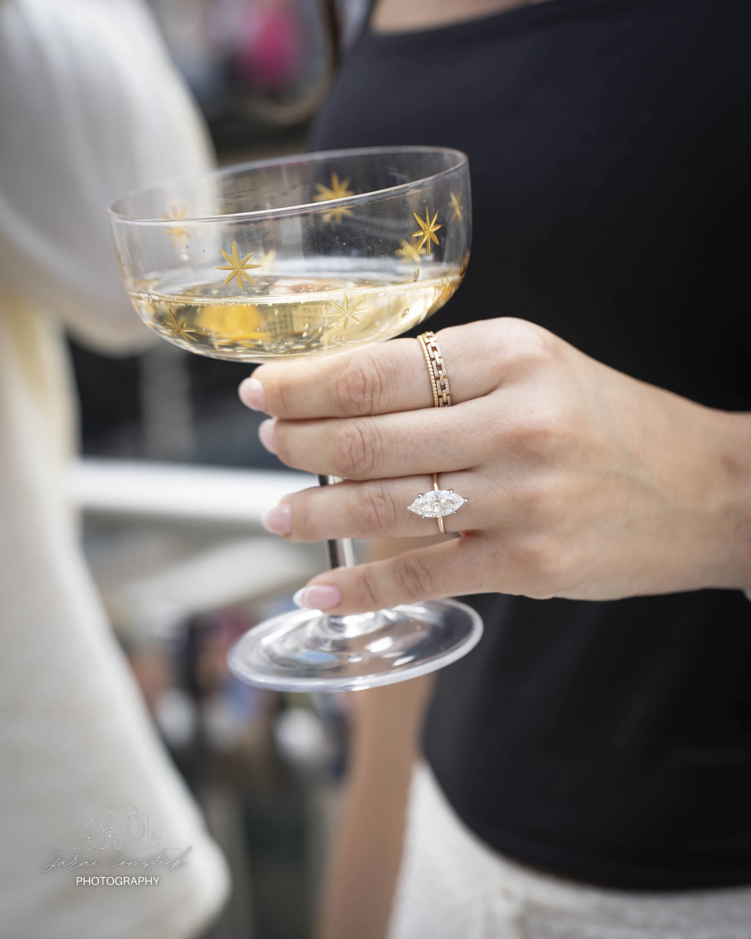 Close-up of a woman holding a clear glass champagne coupe with star patterns, filled with a bubbly drink, showing her hand and rings against a blurred background.