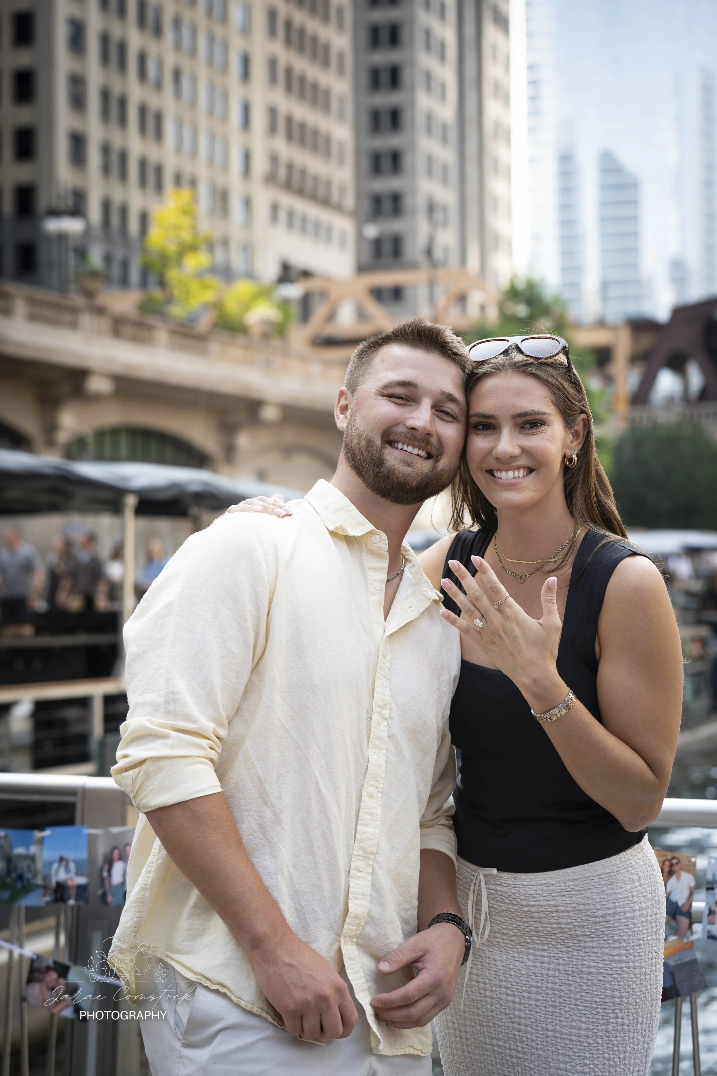 A man and a woman are smiling and posing together outdoors in front of tall buildings and a bridge in an urban setting. The woman is showing her engagement ring.