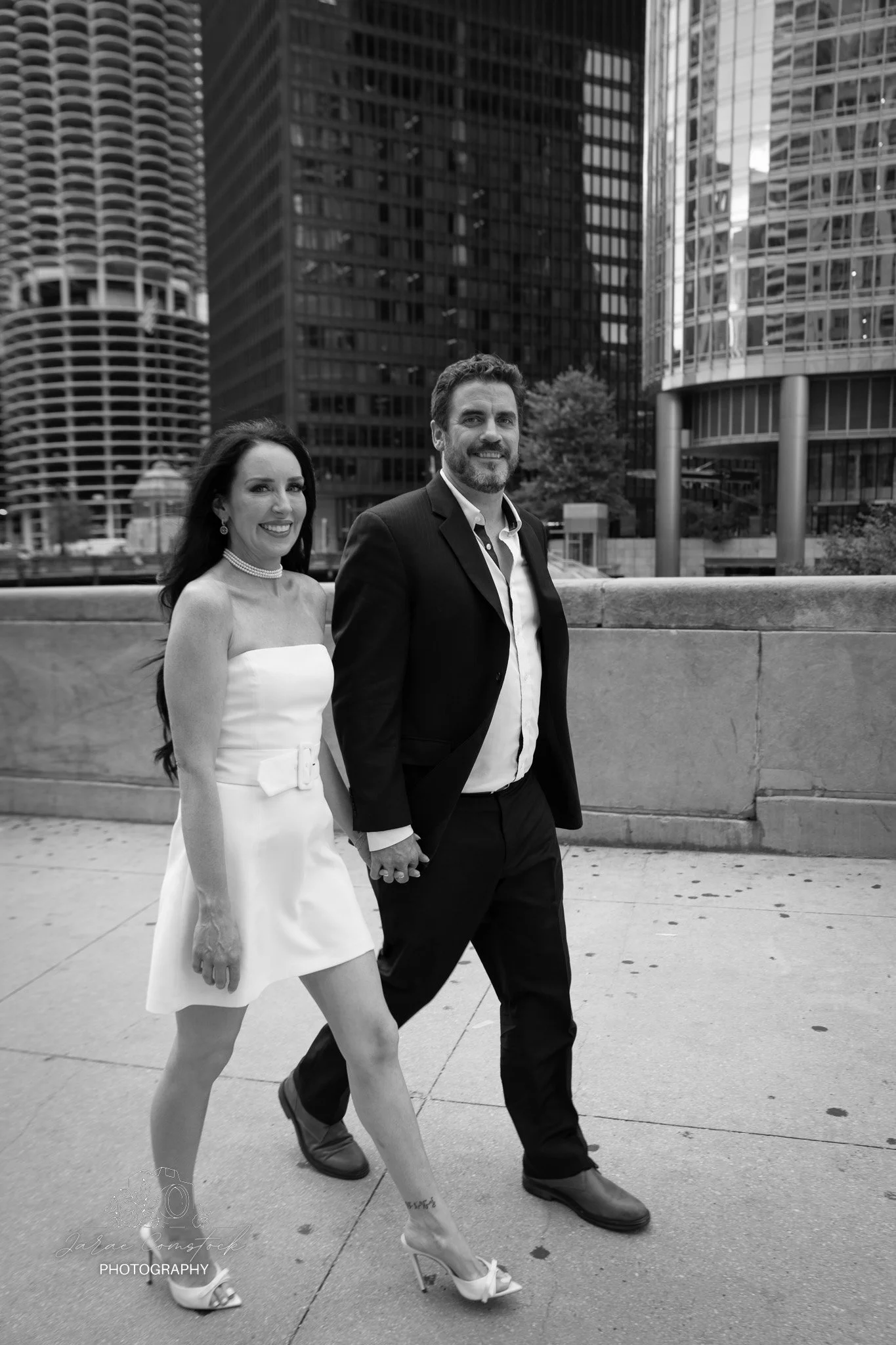 A woman in a white dress and high heels holding hands with a man in a dark suit, walking in an urban area with tall modern buildings in the background.