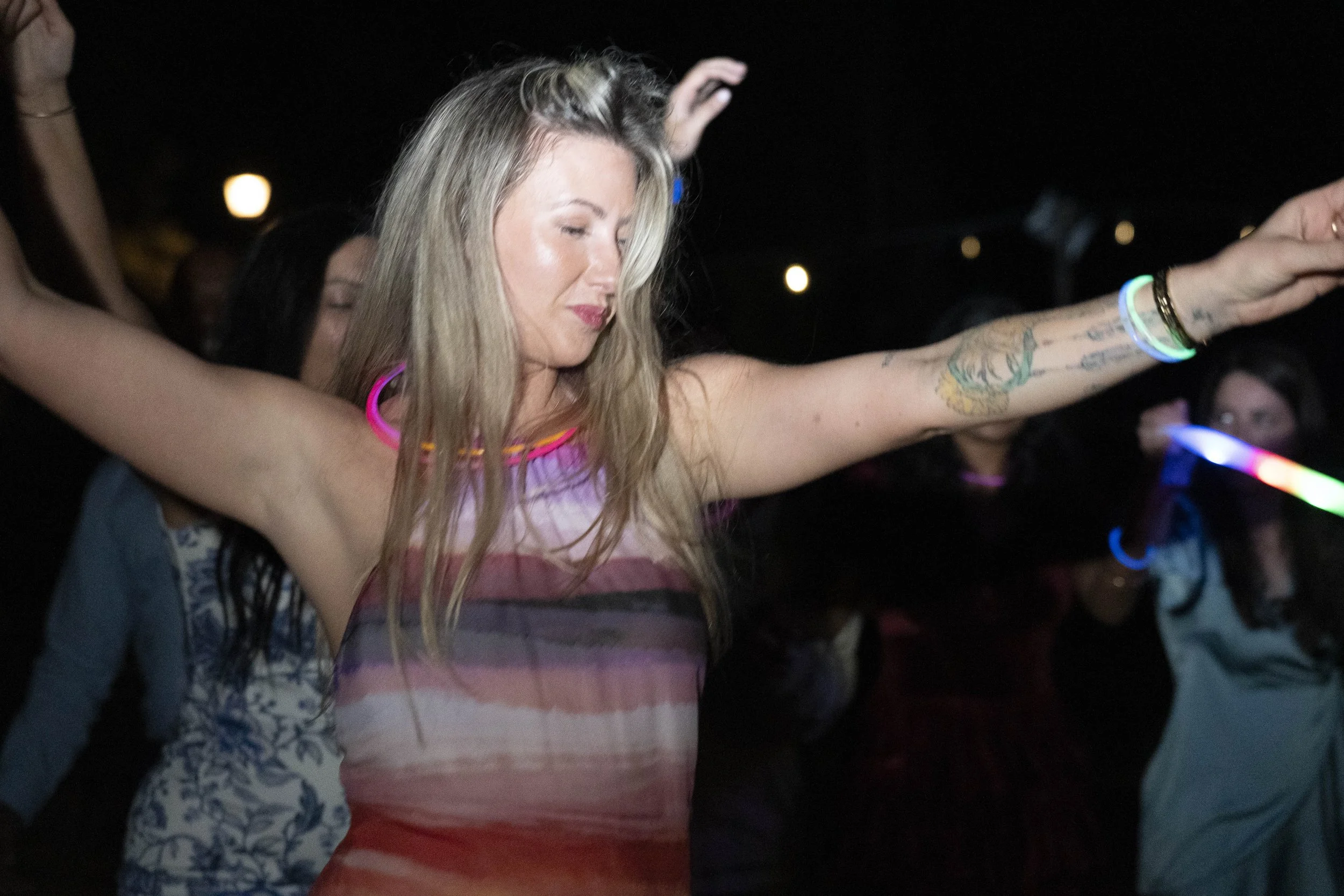 A woman with long blonde hair dancing at a party, wearing a colorful striped top, with glow bracelets on her wrists, in a dark setting with other people in the background.