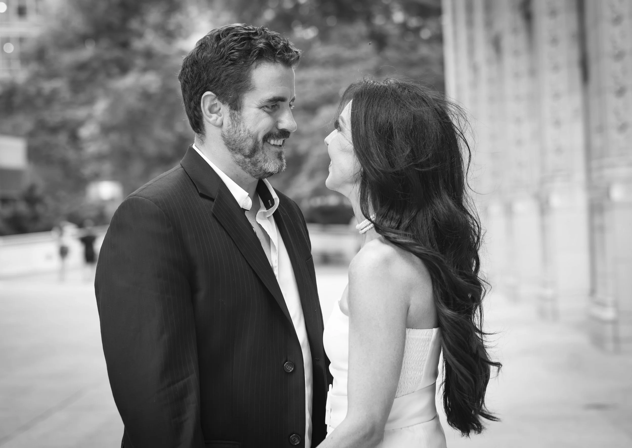 A black and white photo of a man and woman standing close together outdoors, smiling and looking into each other's eyes, with some blurred trees and buildings in the background.