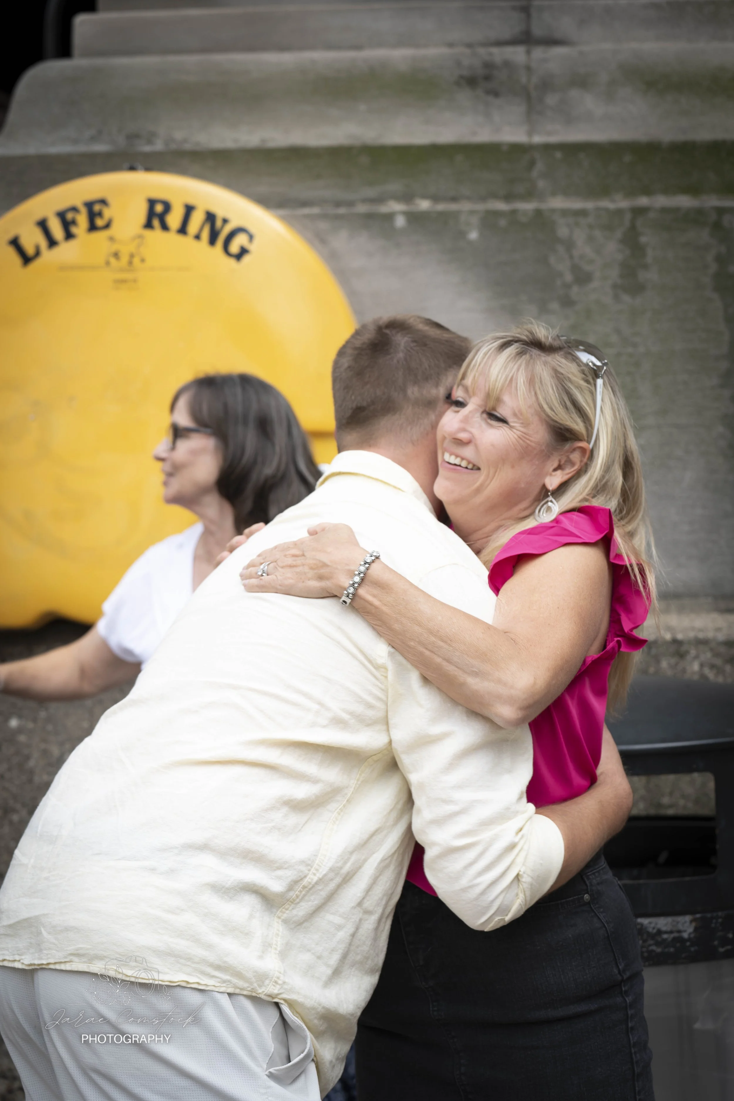 Two women are hugging. One woman is wearing a pink dress with ruffled sleeves and earrings, and has blonde hair. The other woman is wearing a white top and has short hair. In the background, a woman with glasses and dark hair is visible. The scene ta