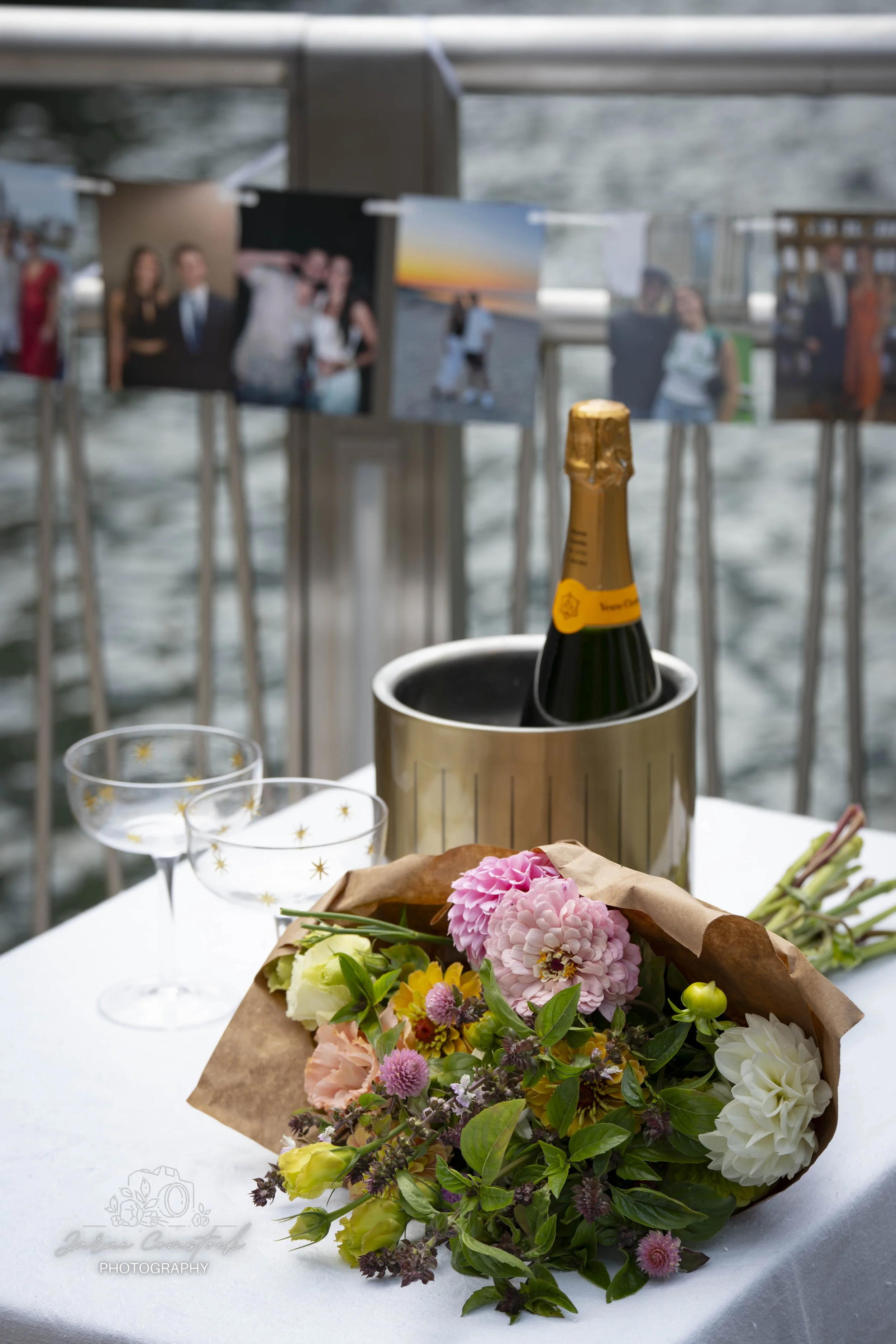 A festive table setting with a bouquet of pink, white, and yellow flowers in brown paper. A bottle of champagne in ice bucket, flutes, and a blurred display of photographs on a balcony overlooking water.