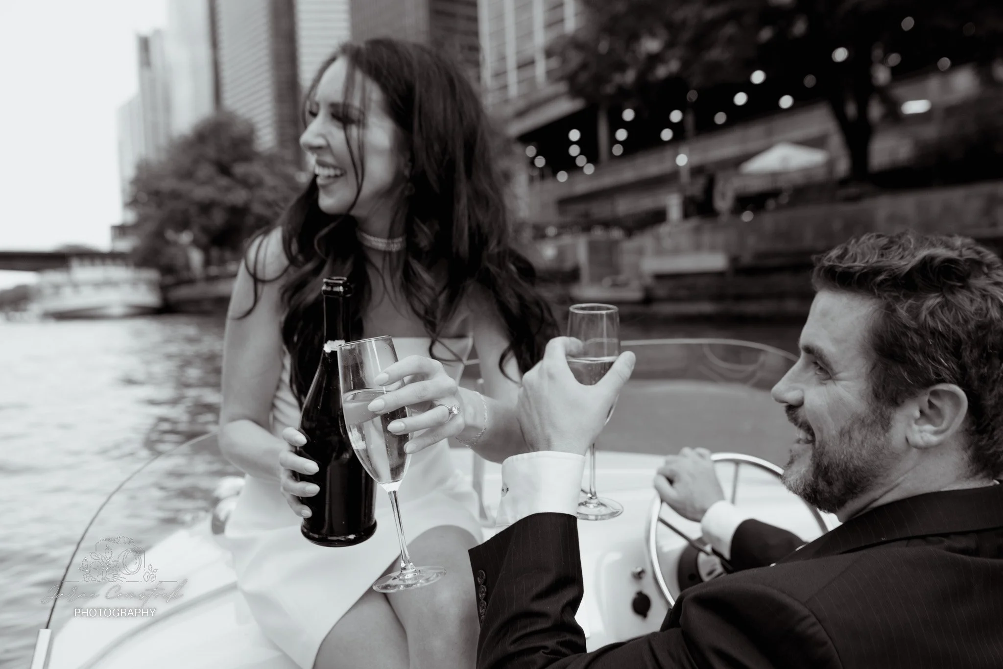 A woman with long dark hair smiling and holding a glass of wine while talking to a man with curly hair and beard, dressed in a suit, who is holding her glass in a toast. They are outdoors near a river with city buildings and a bridge in the backgroun