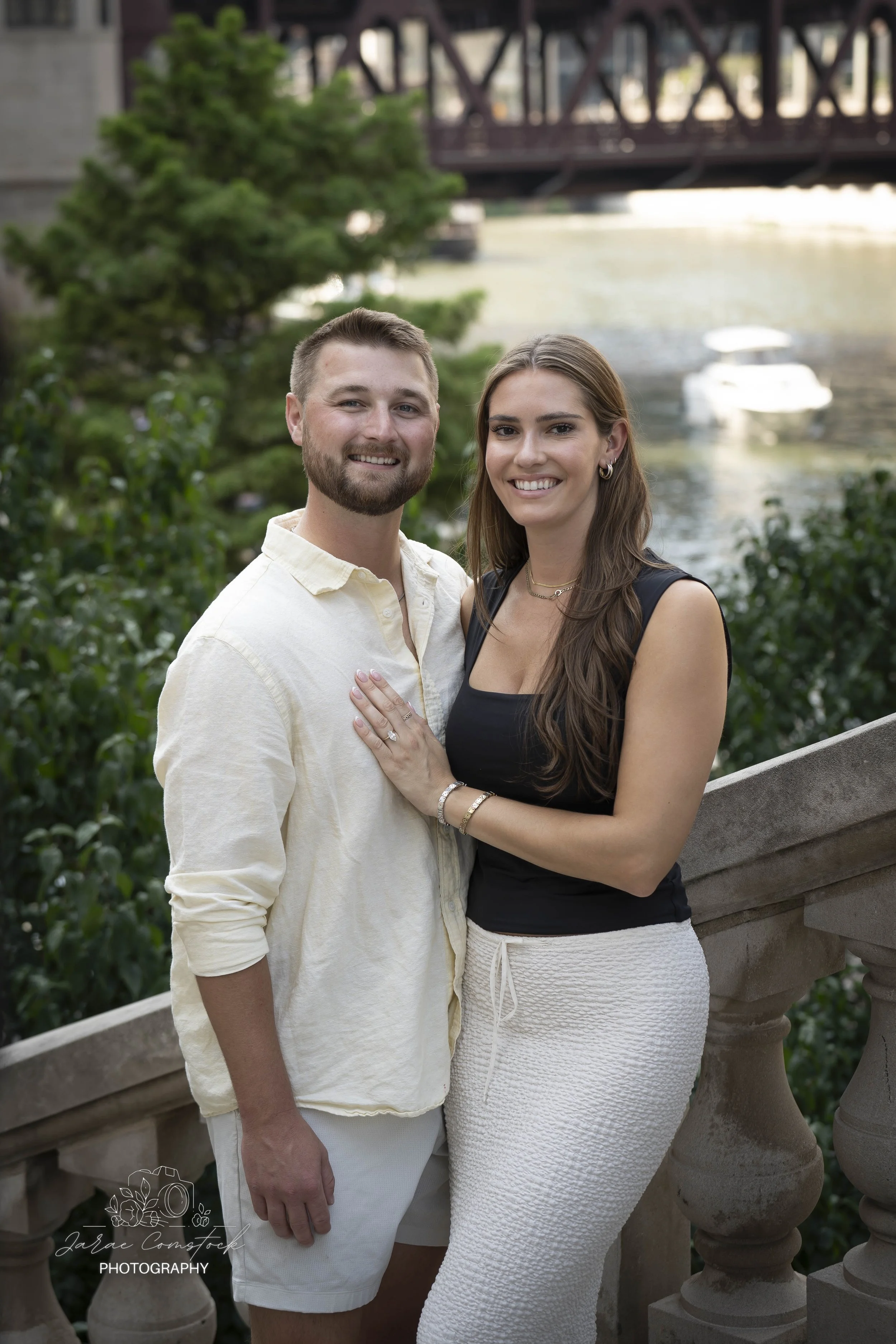 A smiling young man with short hair and a beard wearing a light-colored shirt and shorts, standing next to a smiling young woman with long brown hair wearing a black tank top and white textured skirt, posing outdoors near a river with boats and a bri
