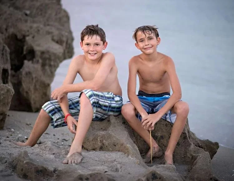 Two boys sitting on rocky shoreline in swim trunks, smiling after playing in the surf