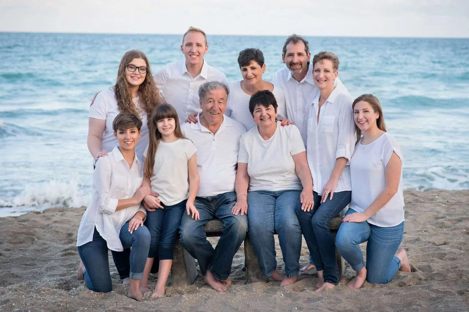 A large extended family photo: everyone in white and jeans, barefoot on a beach near Palm City, Florida with the ocean behind them.