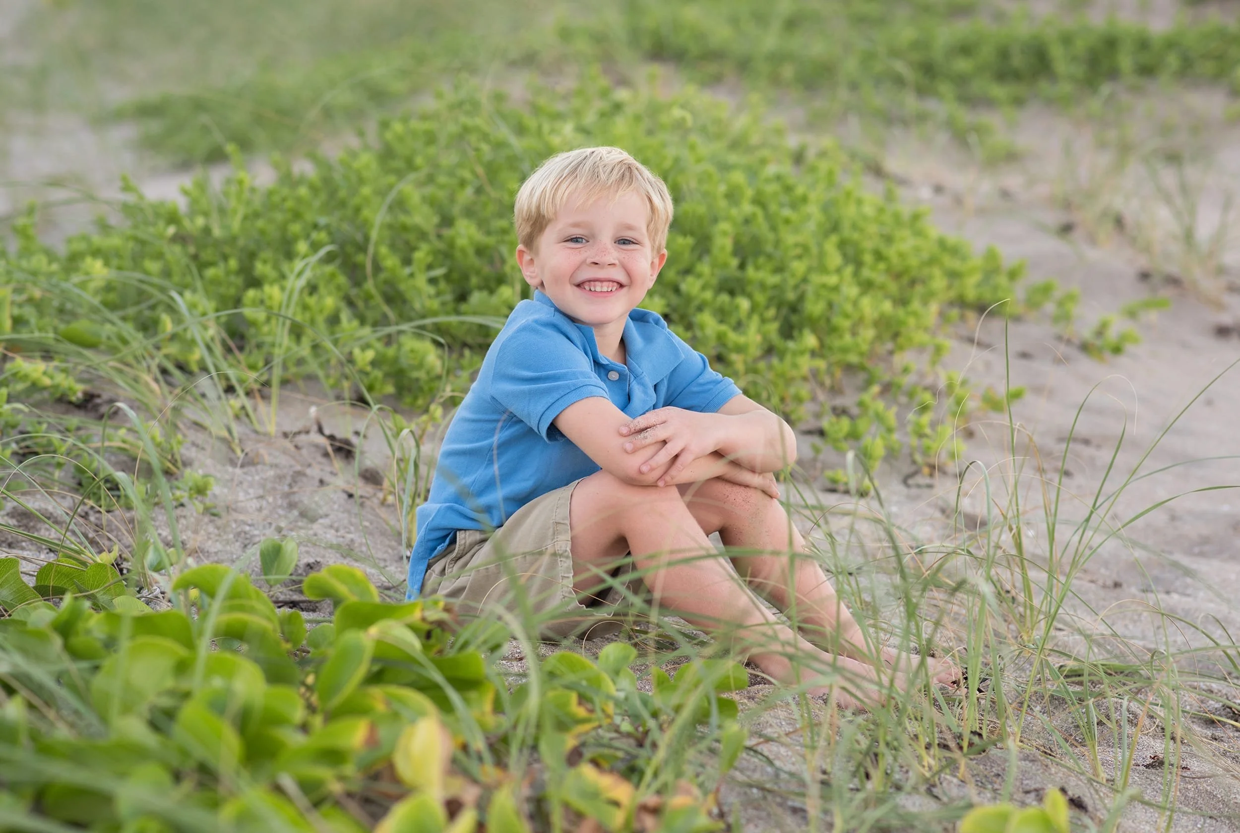 Smiling eight-year-old boy sitting on sandy beach with sea grass in Stuart, Florida during a family session with Dultmeier Photography
