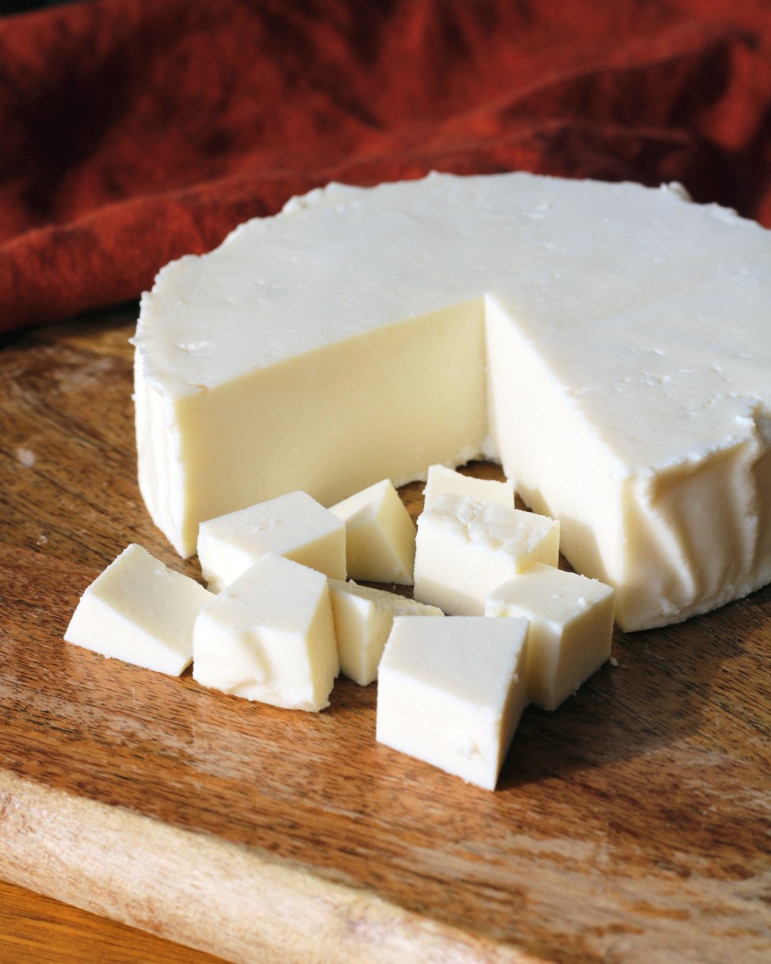 A wheel of white cheese with a slice cut out, placed on a wooden cutting board with cheese cubes in front of it. A red cloth is in the background.