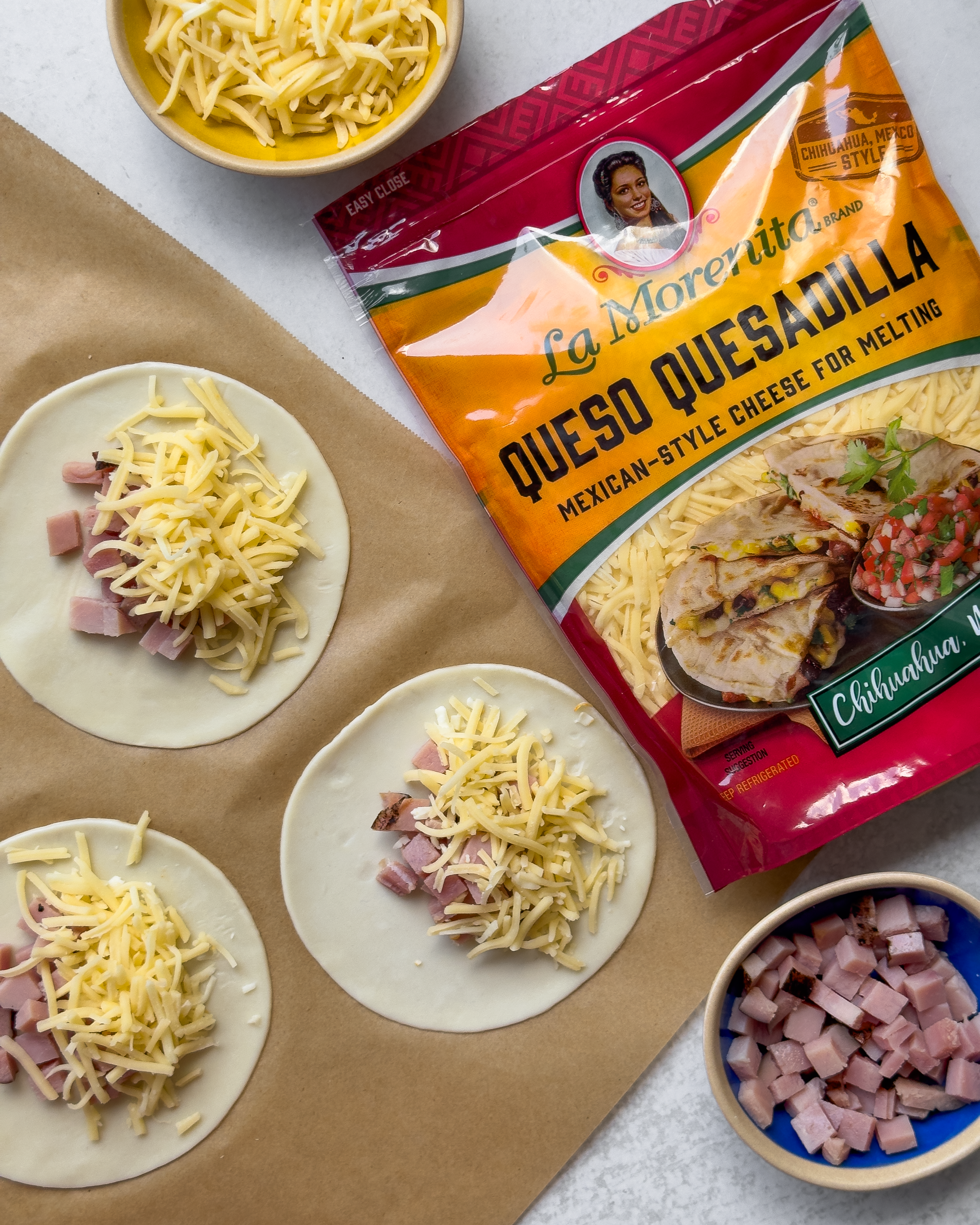 Uncooked Mexican-style cheese quesadillas with shredded cheese, ham, and diced ham on a parchment paper-lined baking sheet, along with a bag of La Morenita queso quesadilla cheese and small bowls of shredded cheese and diced ham.