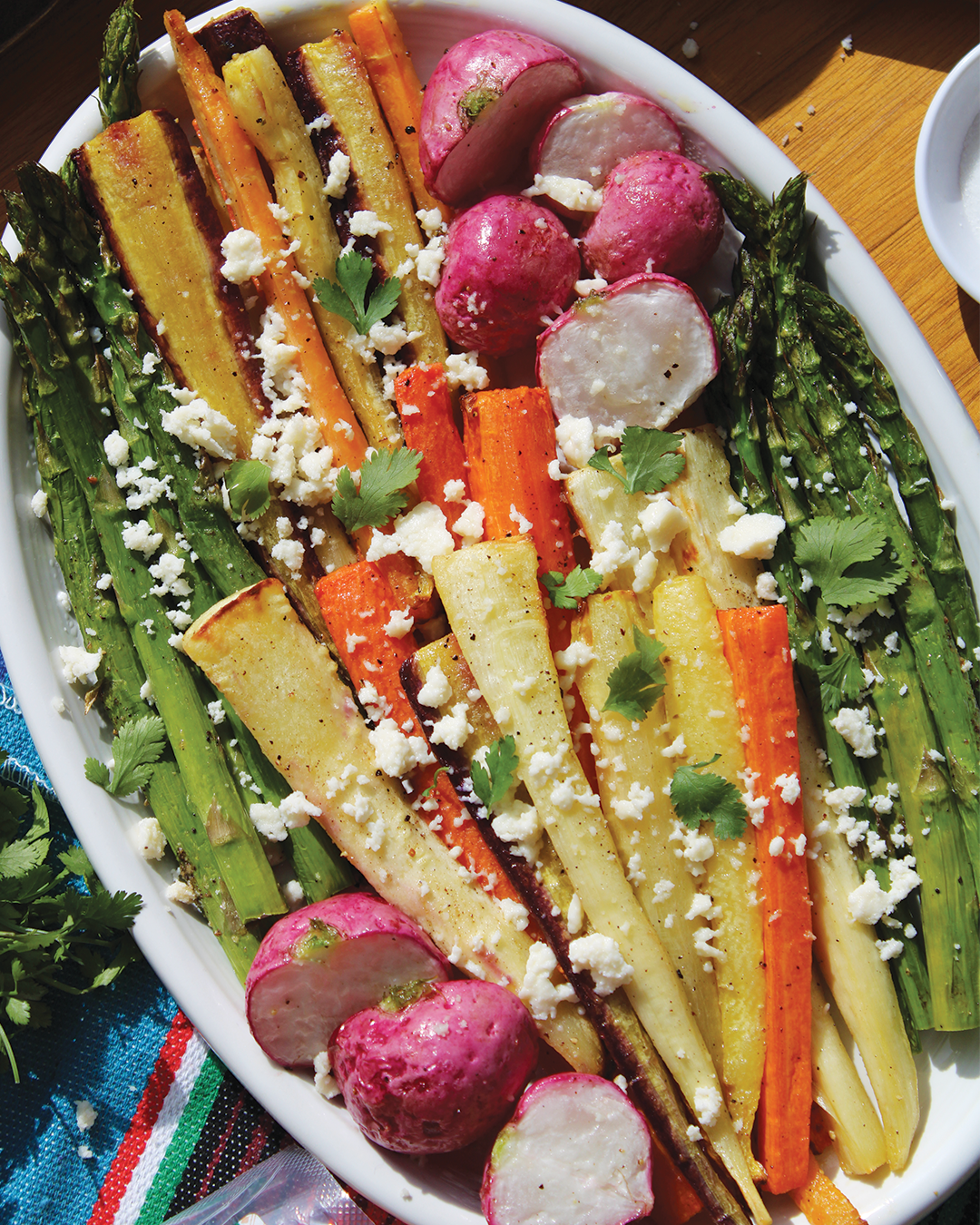 Roasted mixed vegetables including radishes, carrots, parsnips, and asparagus garnished with crumbled cheese and cilantro on a white platter.