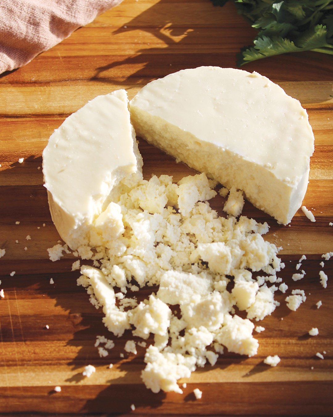 A wheel of crumbled white cheese on a wooden cutting board with green leafy herbs in the background.