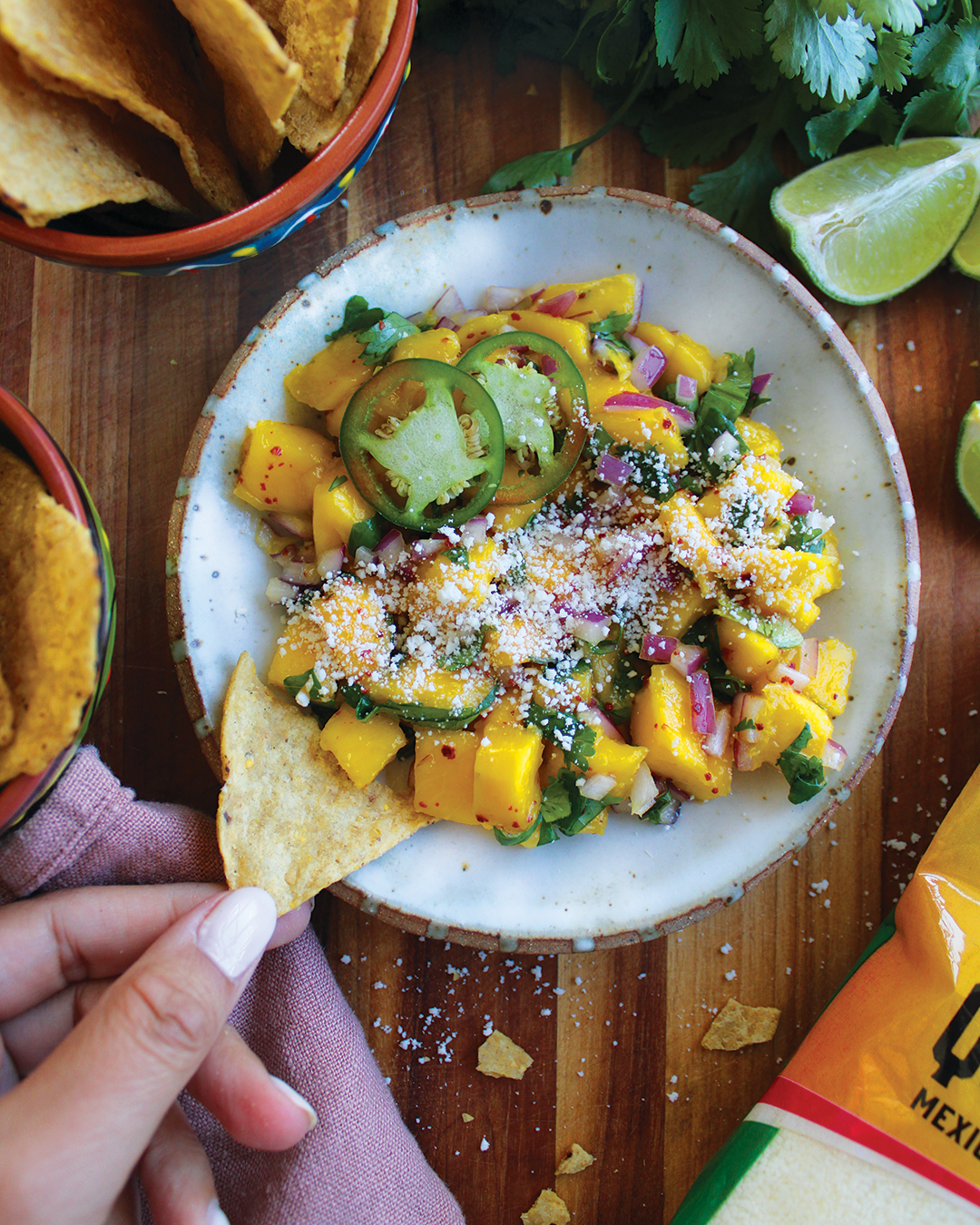 A mango and avocado salad with red onions, sliced jalapeƱos, topped with crumbled cheese, served in a white bowl with tortilla chips on the side, lime wedges nearby, and a hand pulling a tortilla chip from a basket.