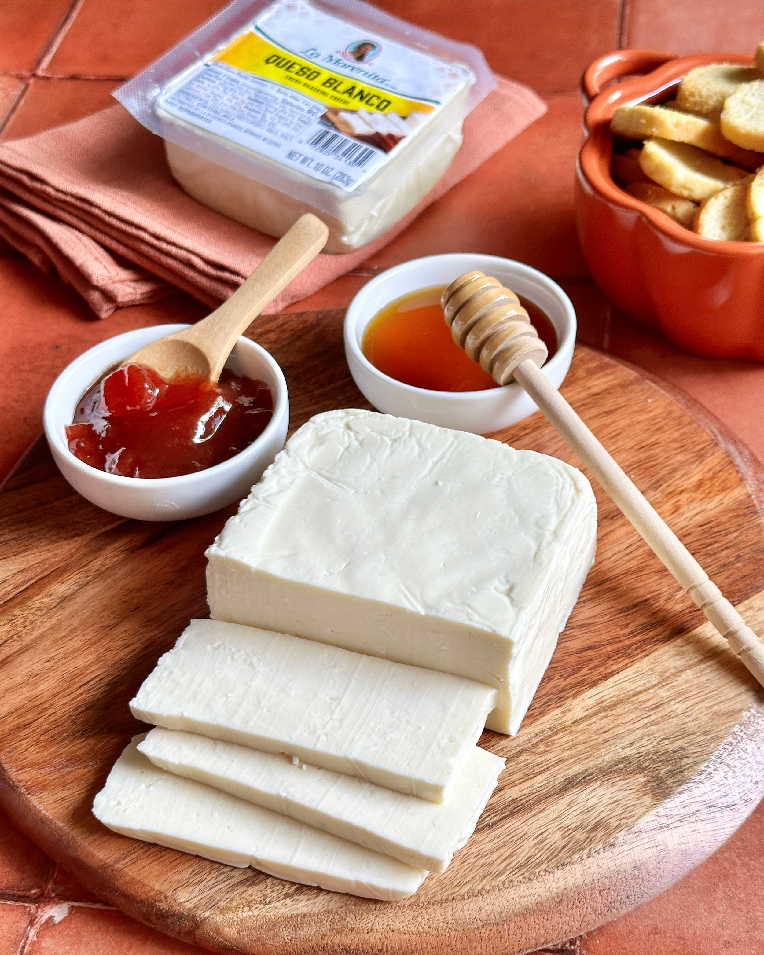 A round wooden board with slices of white cheese, two small bowls with honey and fruit preserves, and a wooden honey dipper. In the background is a package of queso blanco, a bowl of toasted bread slices, and a terracotta dish.