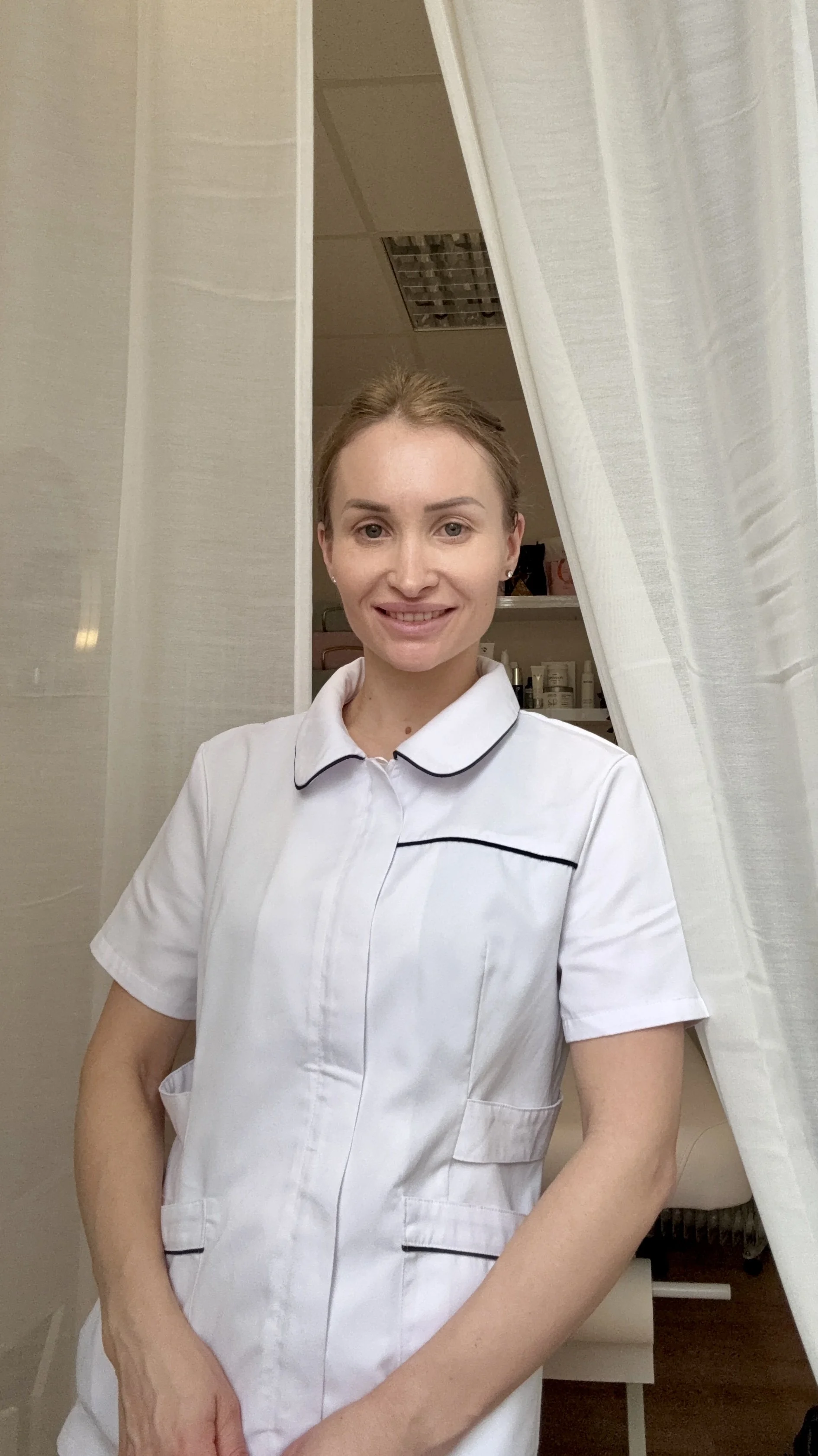 A woman in a white medical uniform standing in a room, smiling at the camera, with shelves and medical supplies in the background.