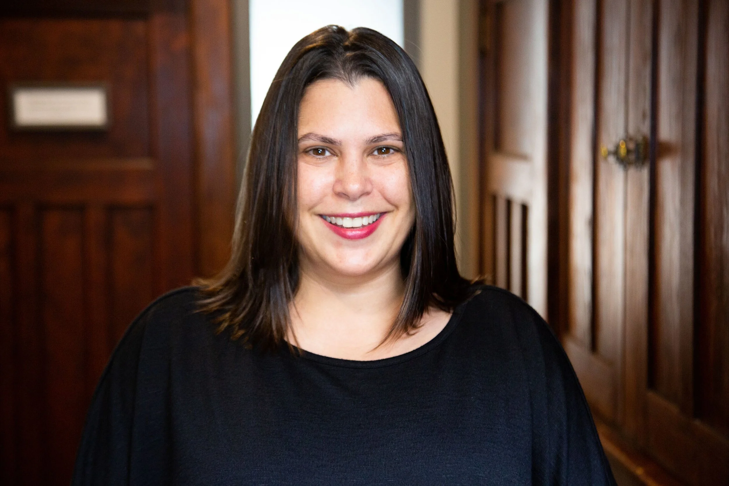 A woman with shoulder-length dark hair and a black top smiling indoors with wooden walls in the background.